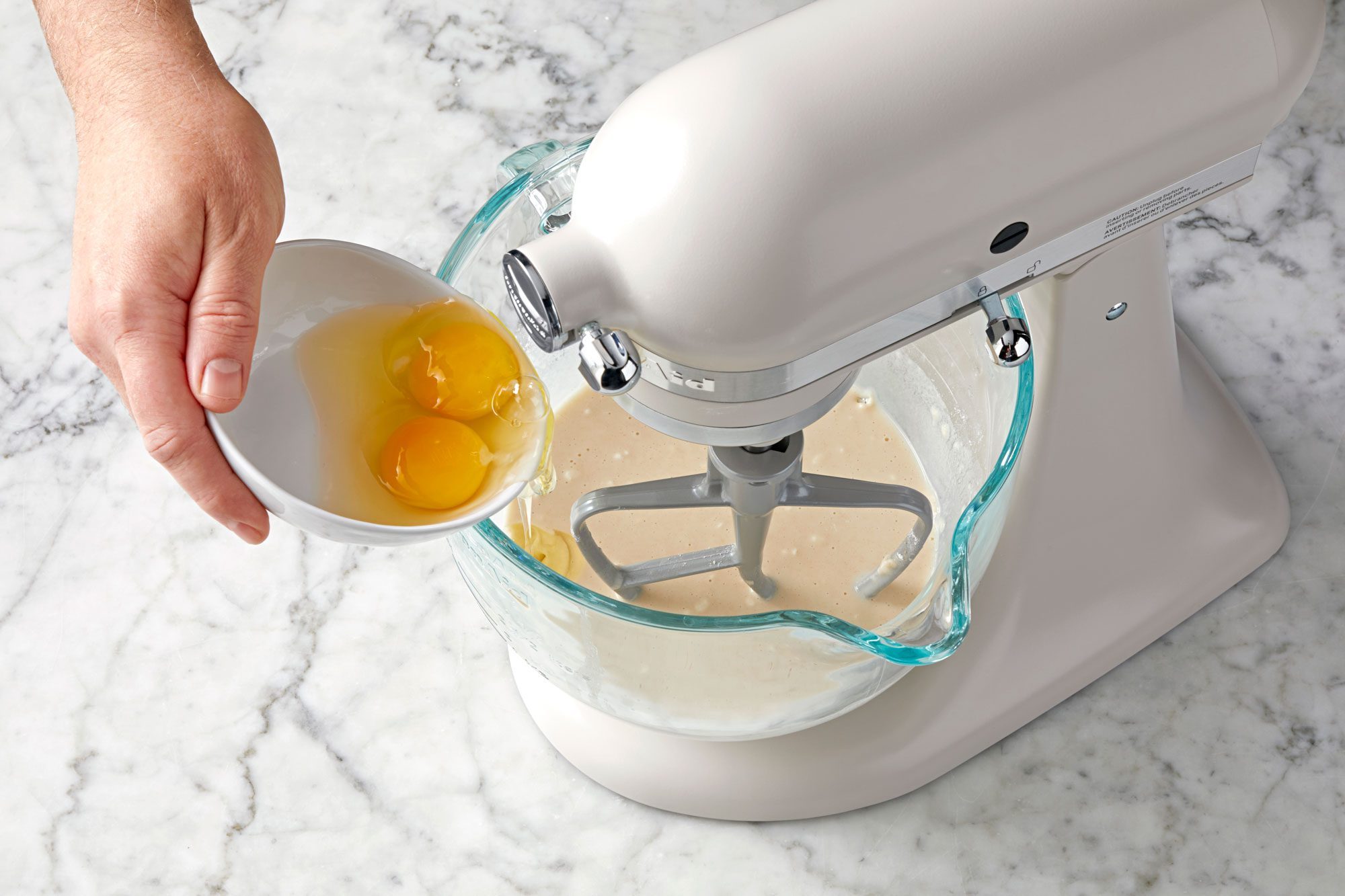 wide shot of pouring two eggs into a glass bowl; stand mixer; marble surface;