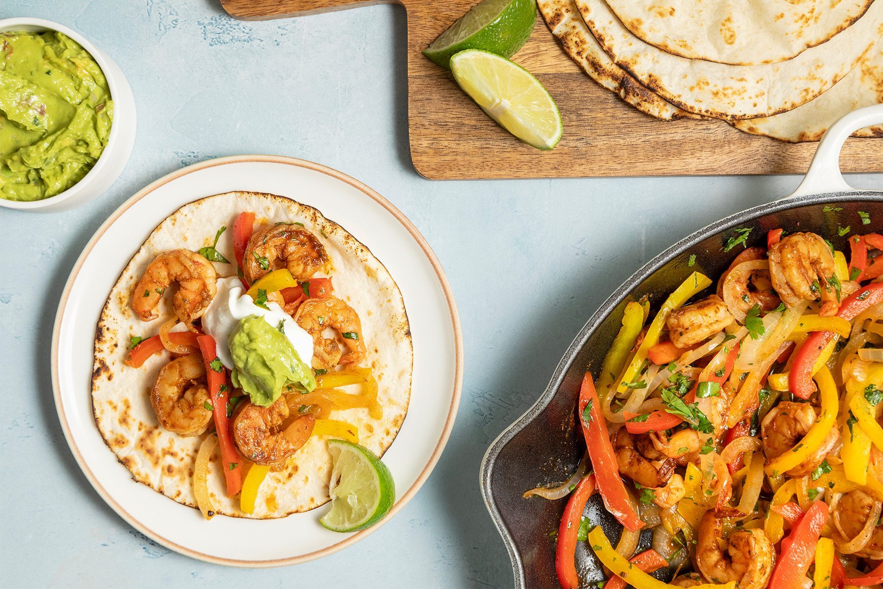 A top view of a shrimp fajita setup. A fajita with shrimp, bell peppers, sour cream, and guacamole on a plate is visible. 