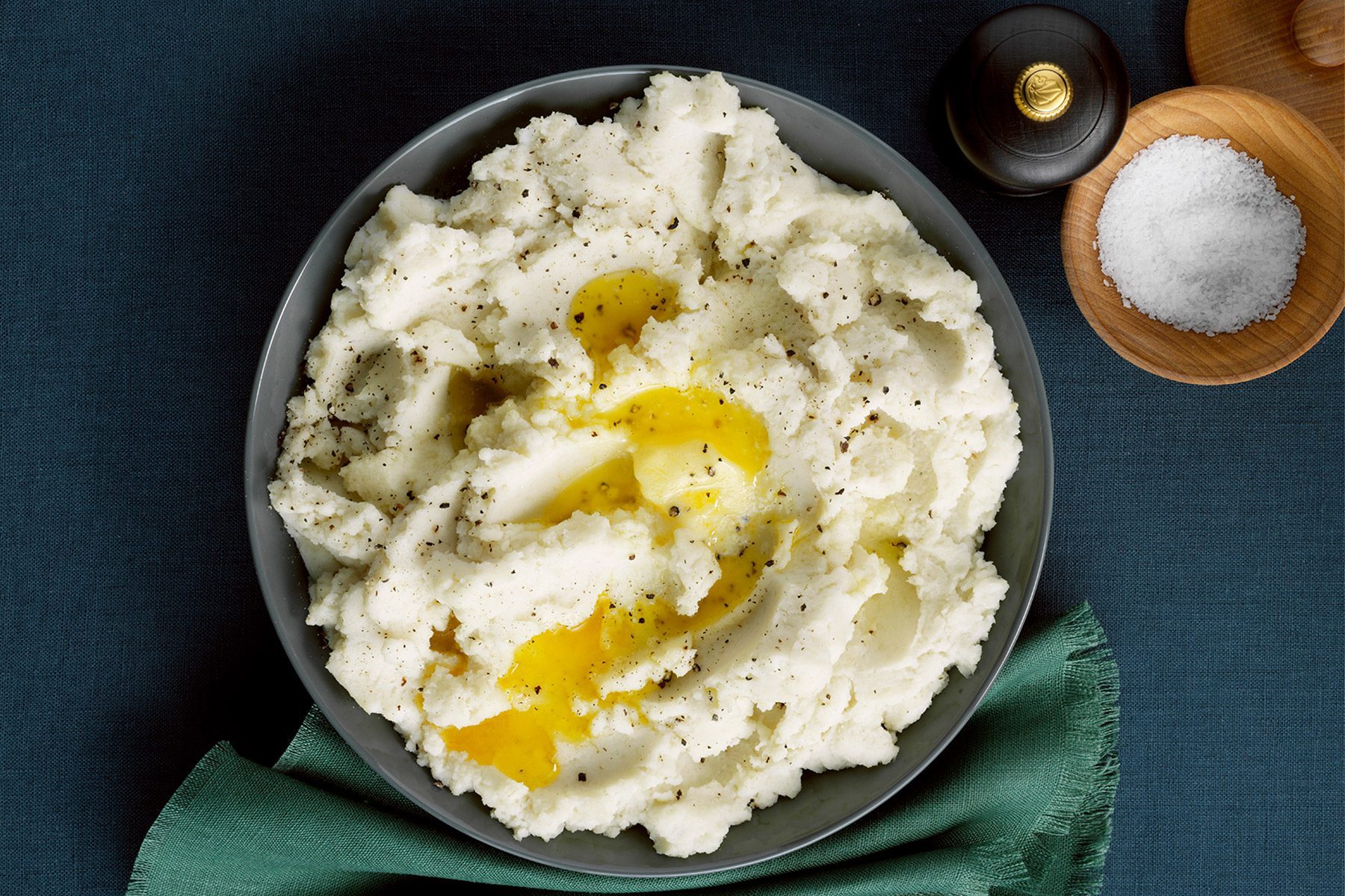 A bowl of creamy mashed potatoes topped with melted butter and sprinkled with black pepper. The bowl is resting on a dark green napkin, and next to it are small wooden bowls containing salt and a pepper grinder on a dark blue tablecloth.