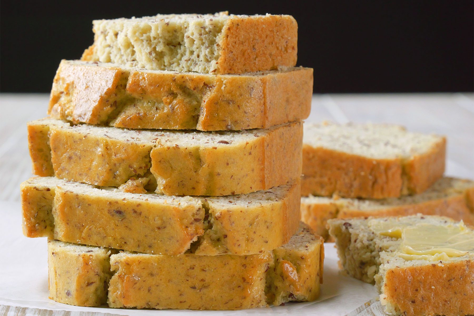A neatly stacked pile of several slices of moist, homemade banana bread with a golden-brown crust and speckled interior, suggesting the presence of nuts or seeds, on a light-colored plate. A partially buttered slice rests beside the stack.