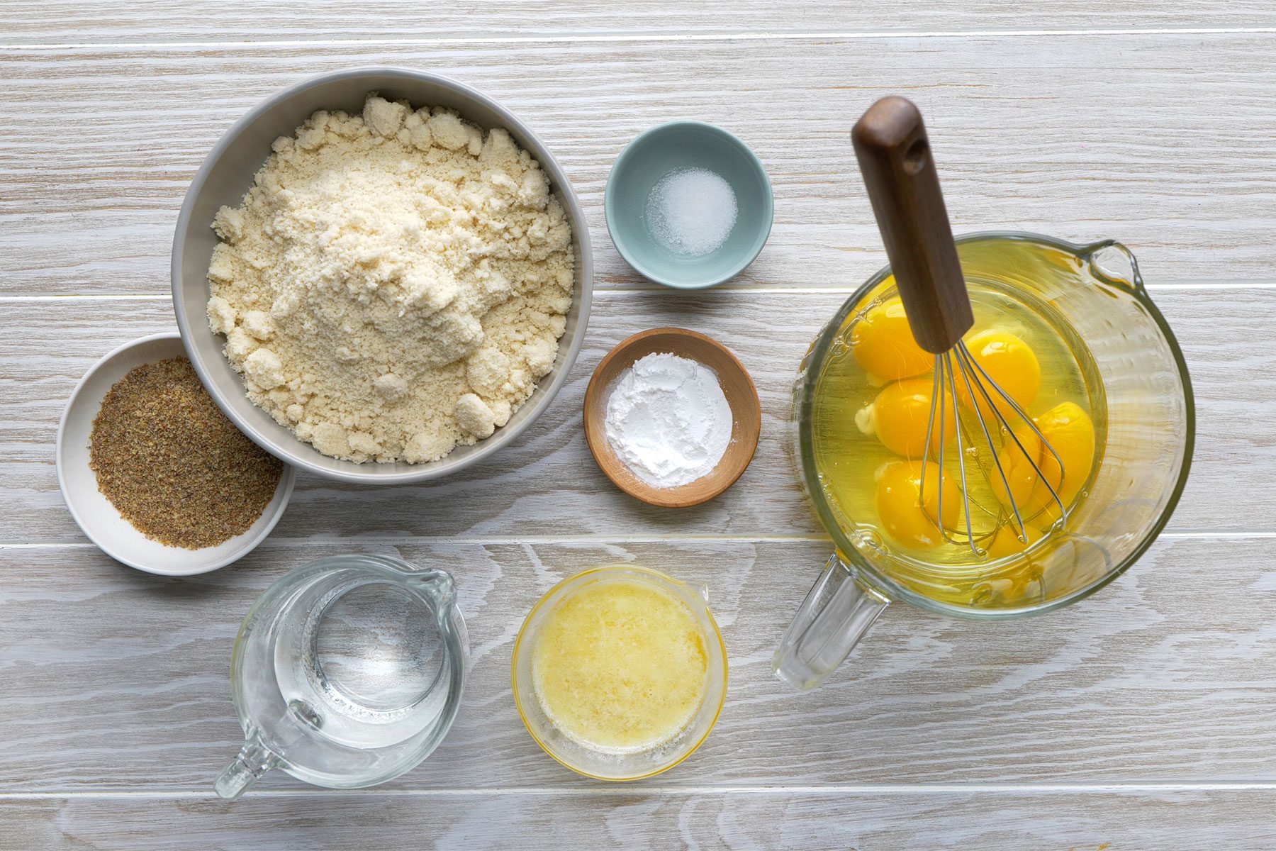 Top-down view of baking ingredients on a wooden surface, including a bowl of flour, a small bowl of salt, a bowl of eggs being whisked, a bowl of baking powder, ground flax seeds, a glass jug of water, and a container of melted butter.