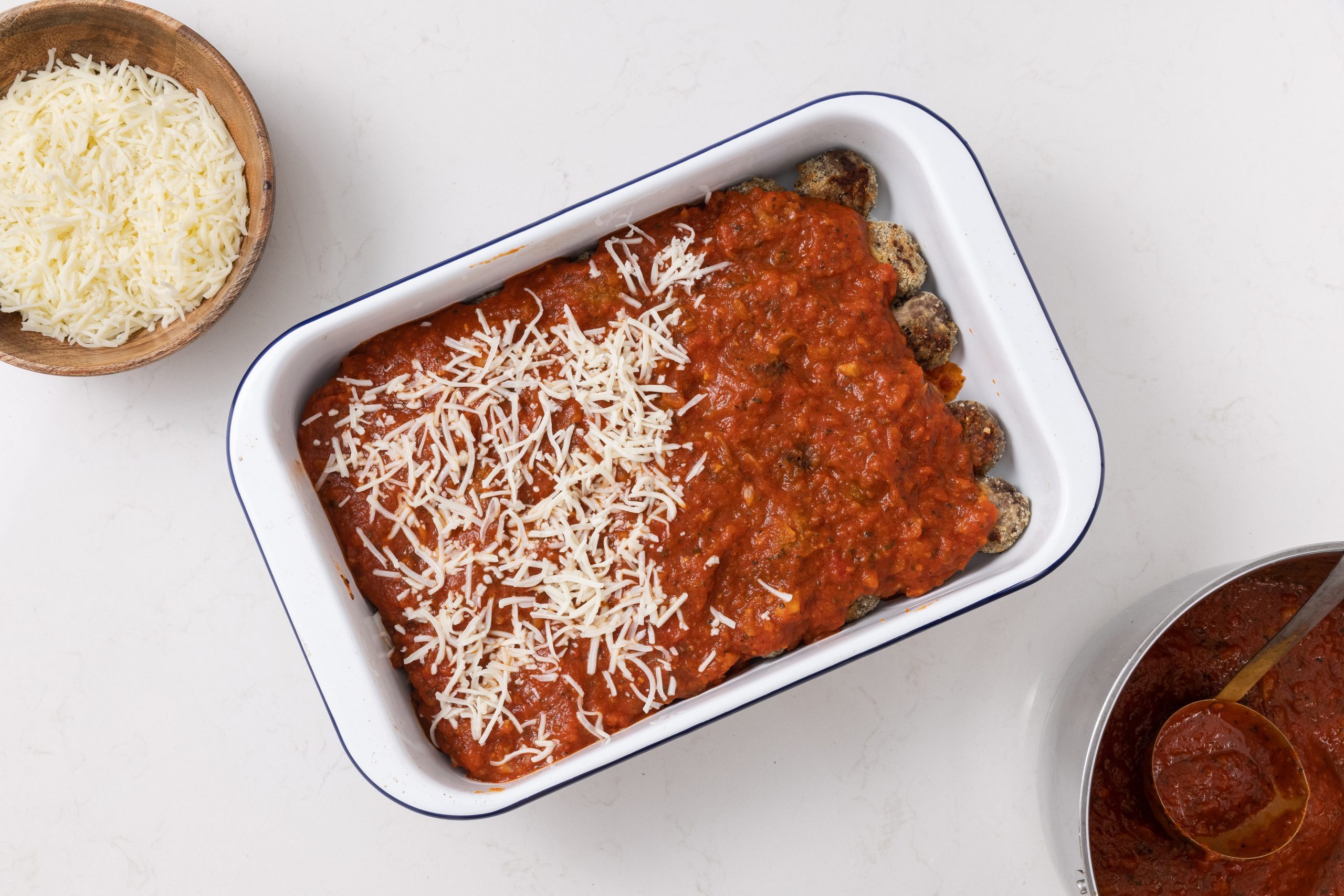 Baked meatballs being layered with tomato sauce and mozzarella in baking dish.