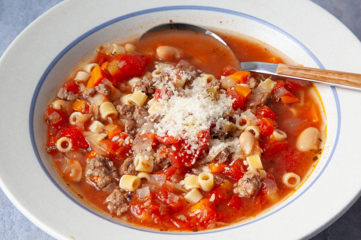 Single servings of Taste of Home Crockpot Pasta Fagioli is white bowls on blue gray surface with small bowl of parmesan cheese