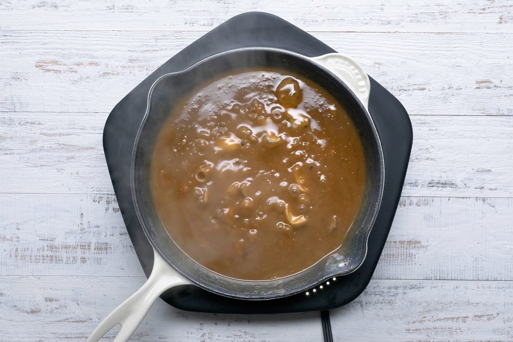 A white cast-iron pan sits on a black trivet, filled with steaming hot brown stew. The stew appears thick with visible chunks, likely of meat or vegetables, floating in the rich sauce. The background is a white wooden surface.