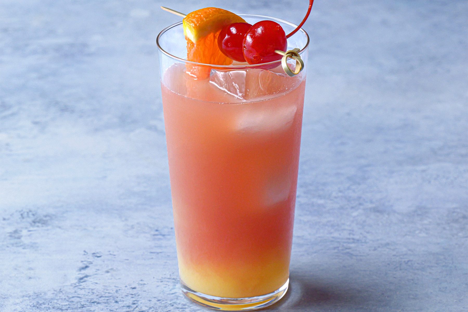horizontal shot; blue textured background; sex on the beach served in glass, garnished with orange wedge and maraschino cherry
