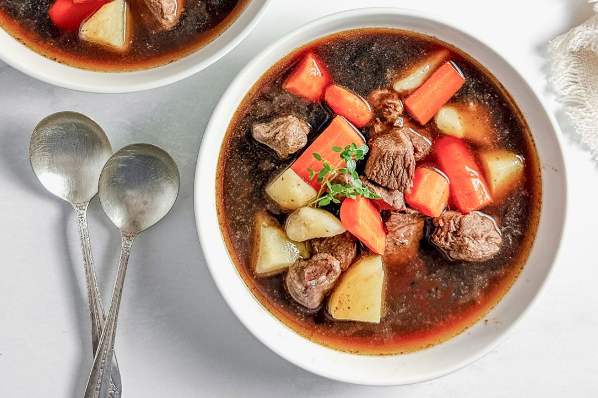 Taste of Home venison stew in white ceramic bowls on a marble surface next to a cloth napkin.