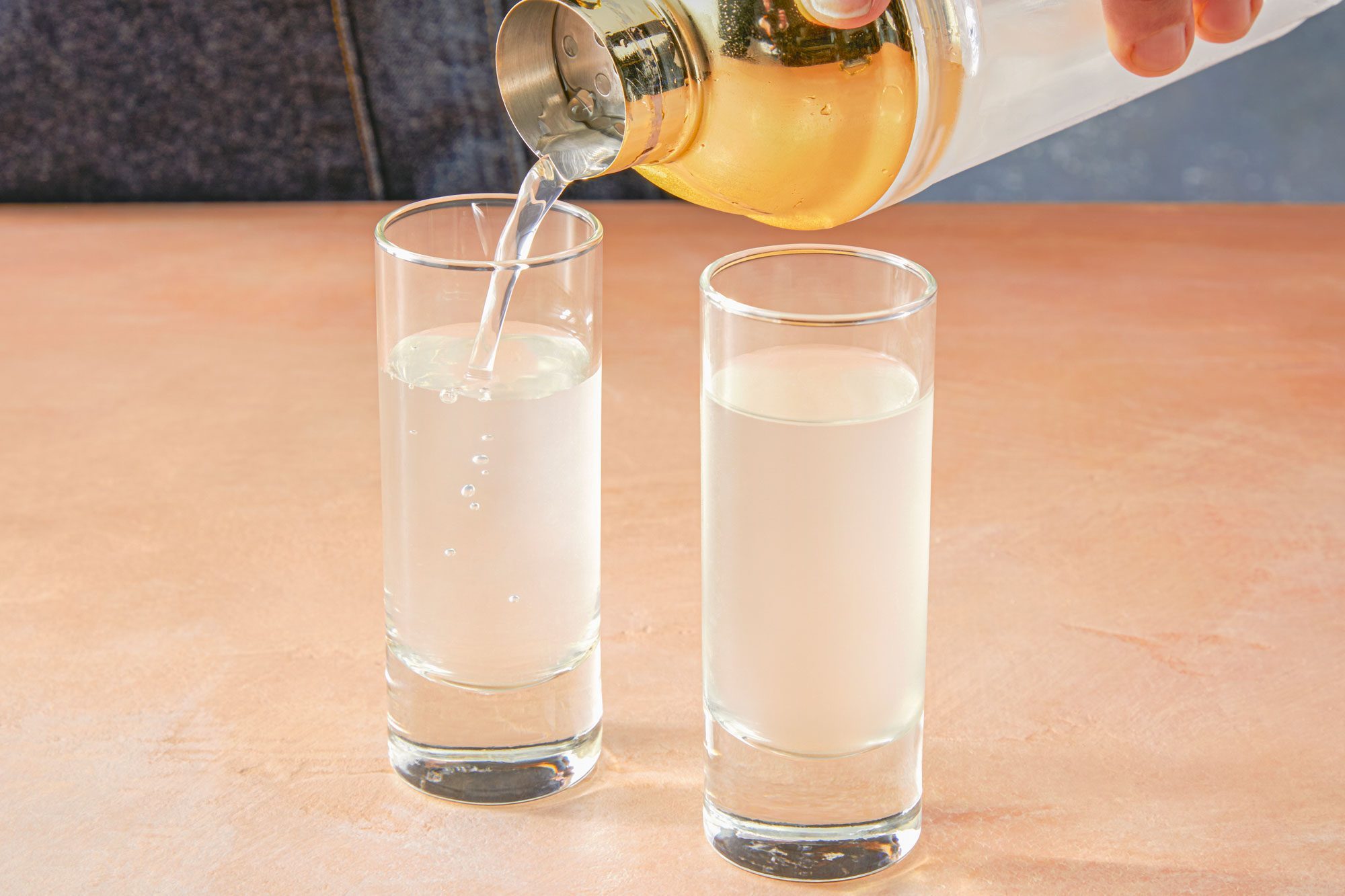 Close shot of pouring into 2 shot glasses; shaker; light orange background;