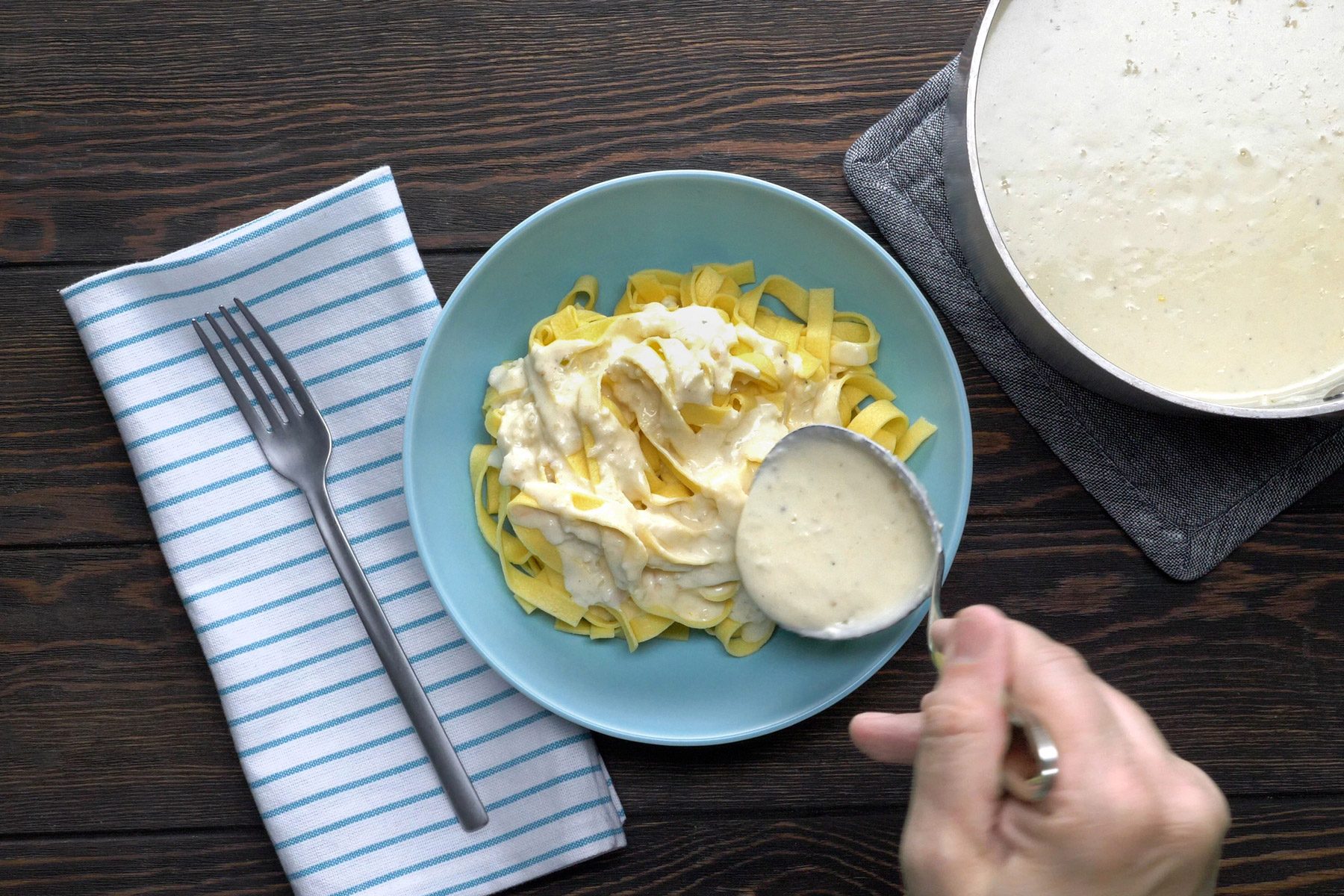 A hand can be seen using a spoon to coat the pasta with Alfredo sauce.