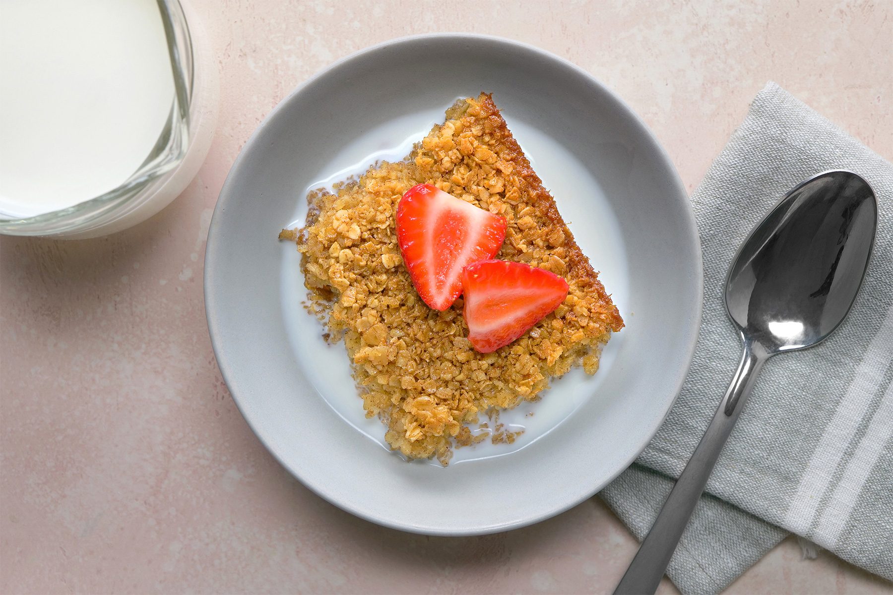 overhead shot; light peach background; Baked Oatmeal served with milk on a white plate with silver spoon placed over grey kitchen towel, with a jar of milk;