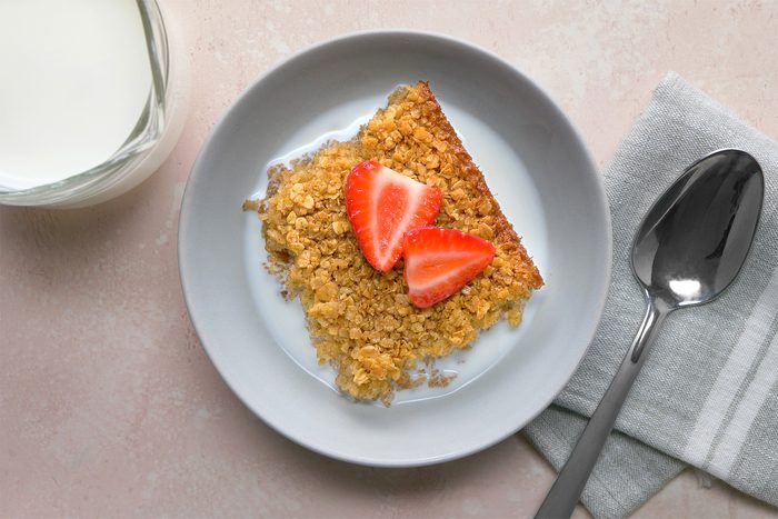 overhead shot; light peach background; Baked Oatmeal served with milk on a white plate with silver spoon placed over grey kitchen towel, with a jar of milk;
