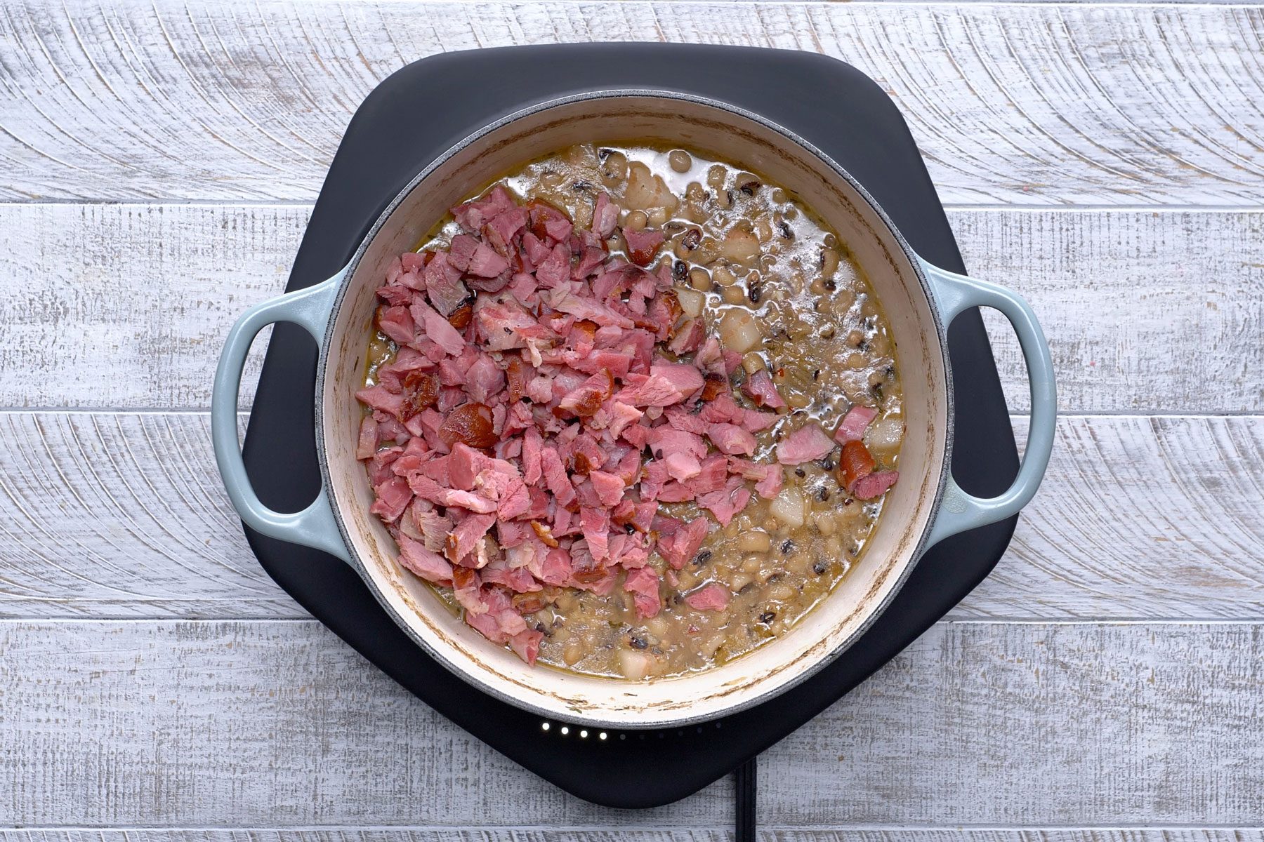 Chopped ham hocks in a broth with peas in a pot on a stovetop against a textured wooden background.
