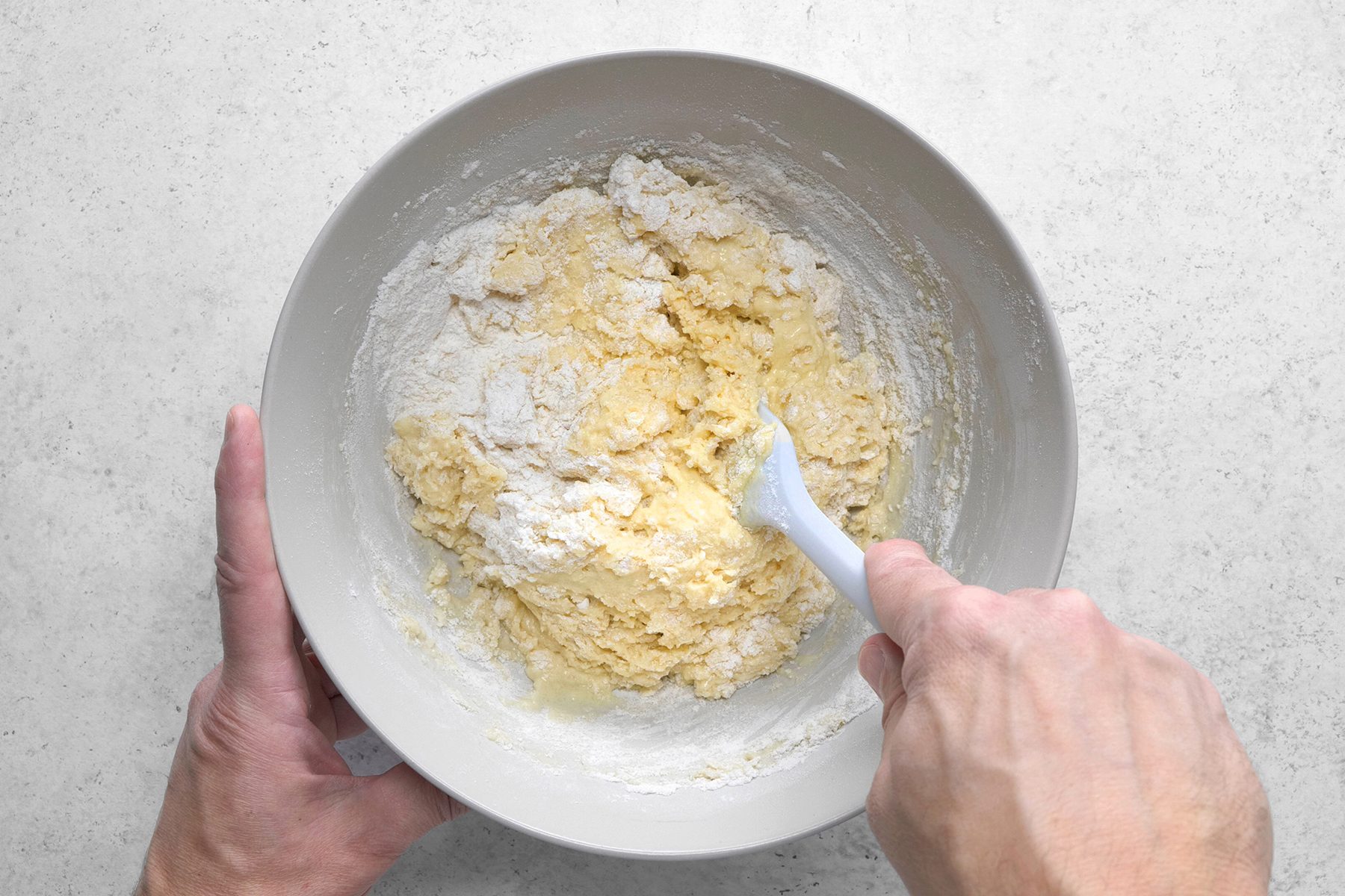 A person is mixing a lumpy dough with a white plastic spatula in a grey bowl. The dough includes visible flour and wet ingredients, and the bowl is placed on a light-colored countertop. Both of the person's hands are partially visible in the image.
