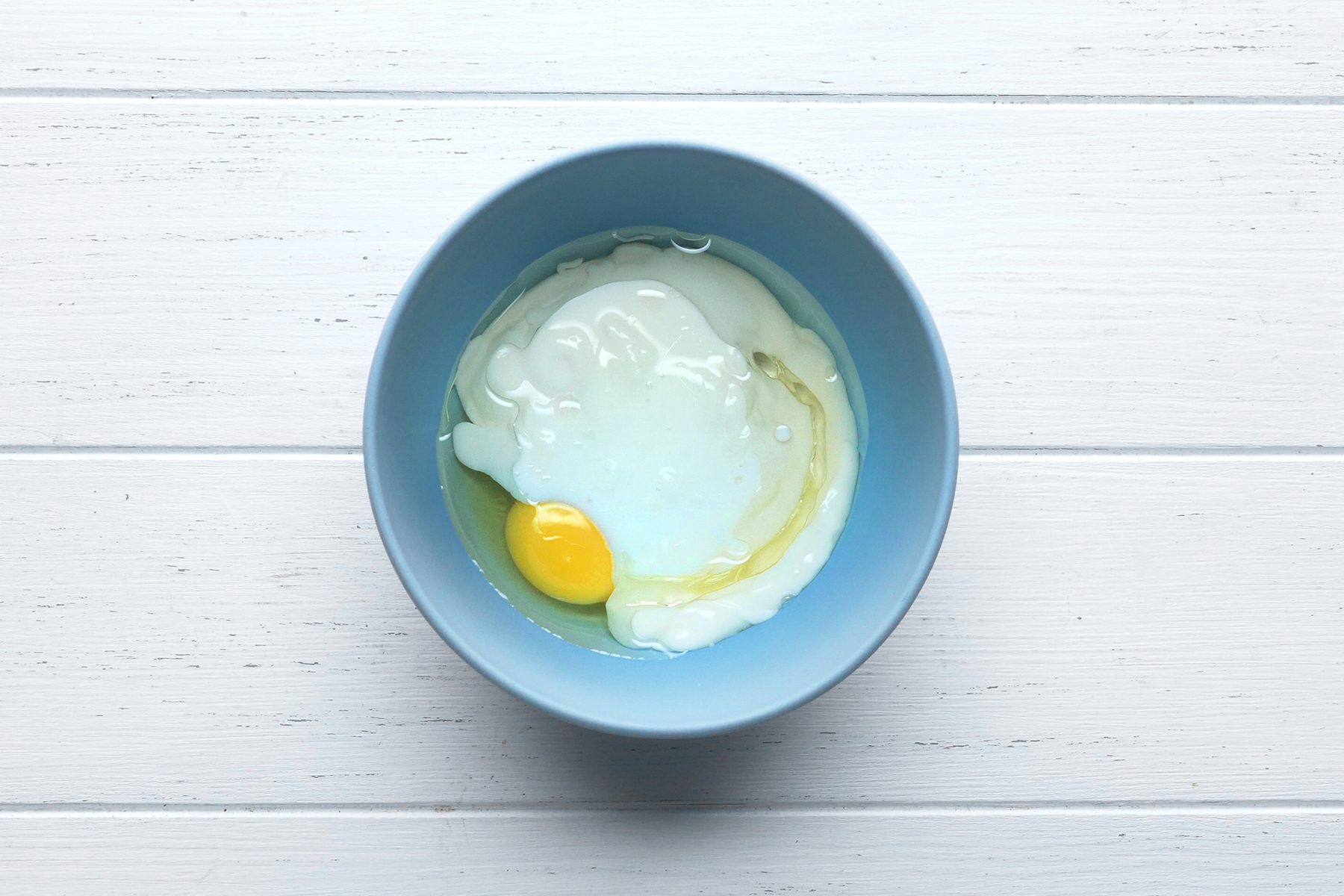 A cracked raw egg with a visible yolk and egg white is in a light blue bowl, set on a white wooden surface. 