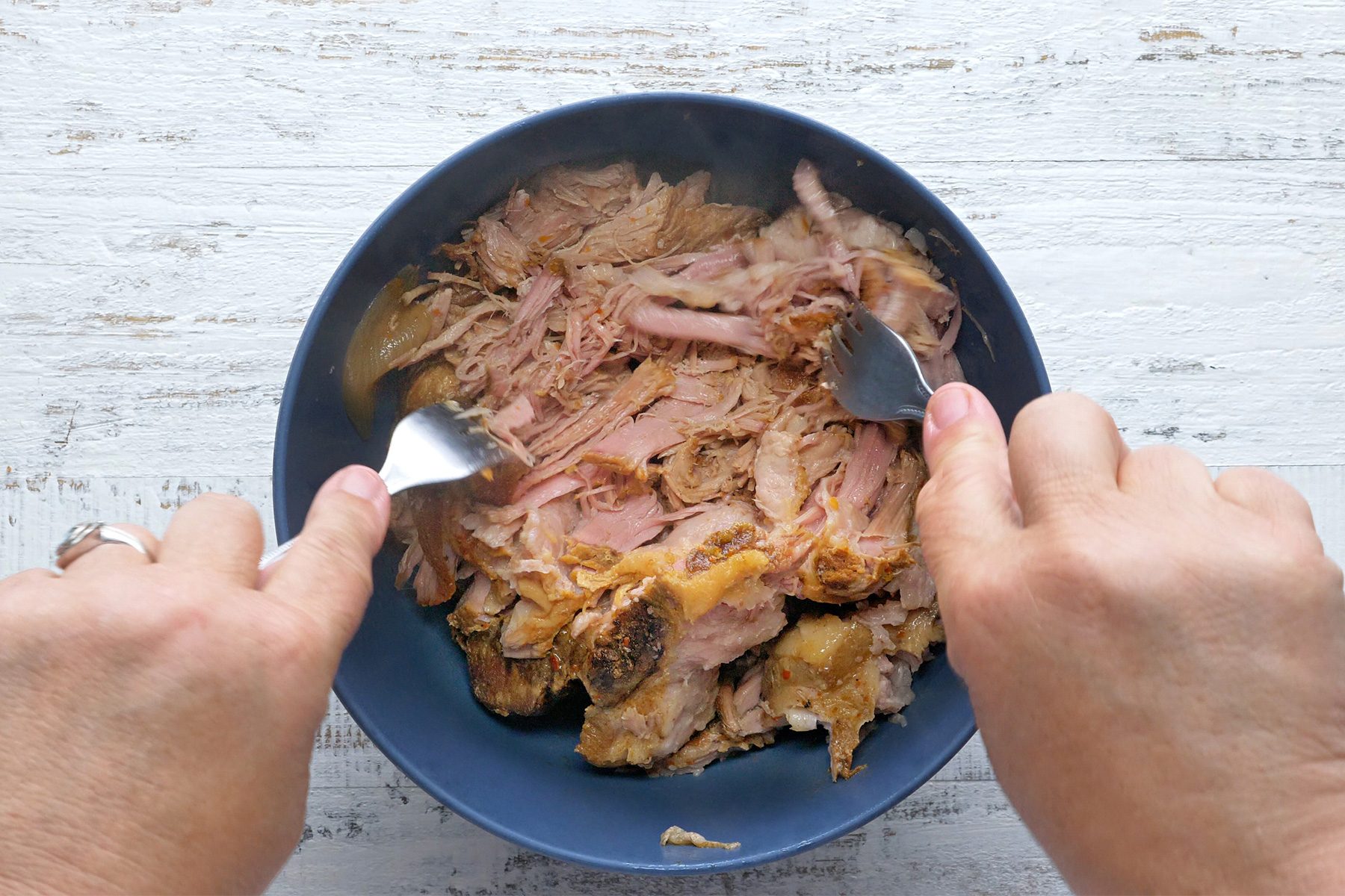 overhead shot; white textured background; Shredding pork with 2 forks in a bowl;