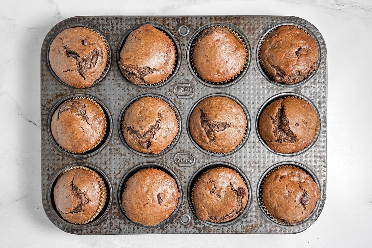 Taste of Home Chocolate Banana Muffins in a metal muffin tin on a marble surface.