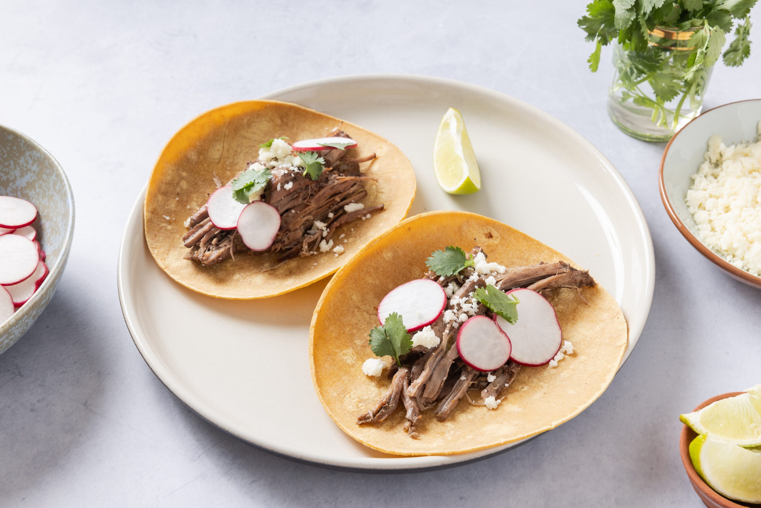 Tacos served on plate with sides of cotija cheese, radishes, lime wedges and cilantro.