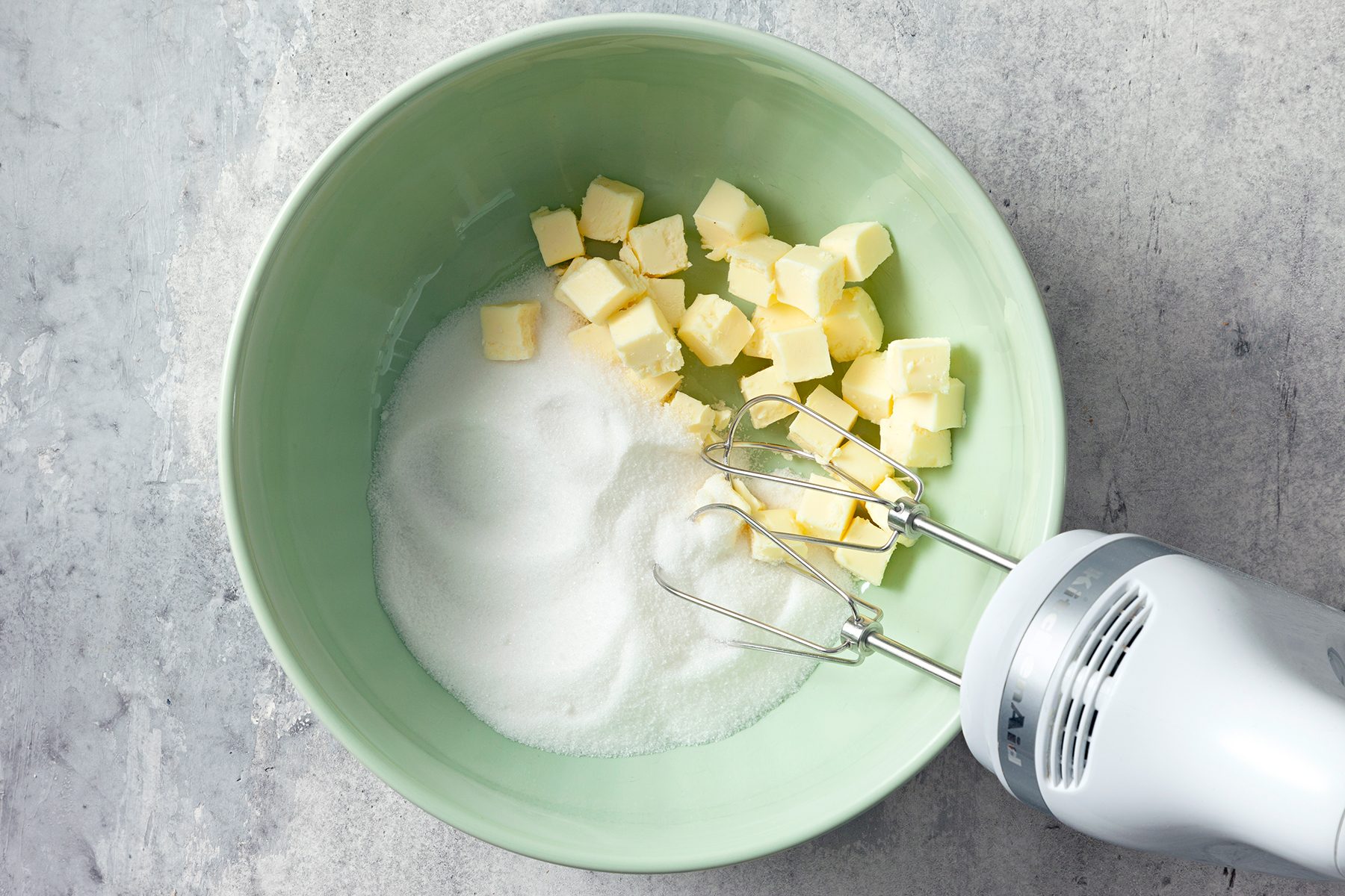 A green mixing bowl contains cubed butter and sugar. An electric hand mixer with beaters is partially submerged in the bowl, ready to mix the ingredients. The background is a gray textured surface.