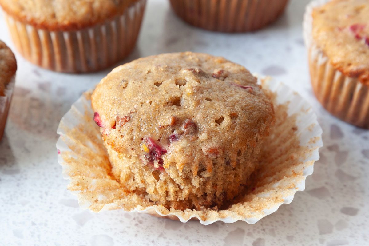 Close up of Taste of Home Cranberry Muffins on a marble surface, one muffin with the liner peeling off