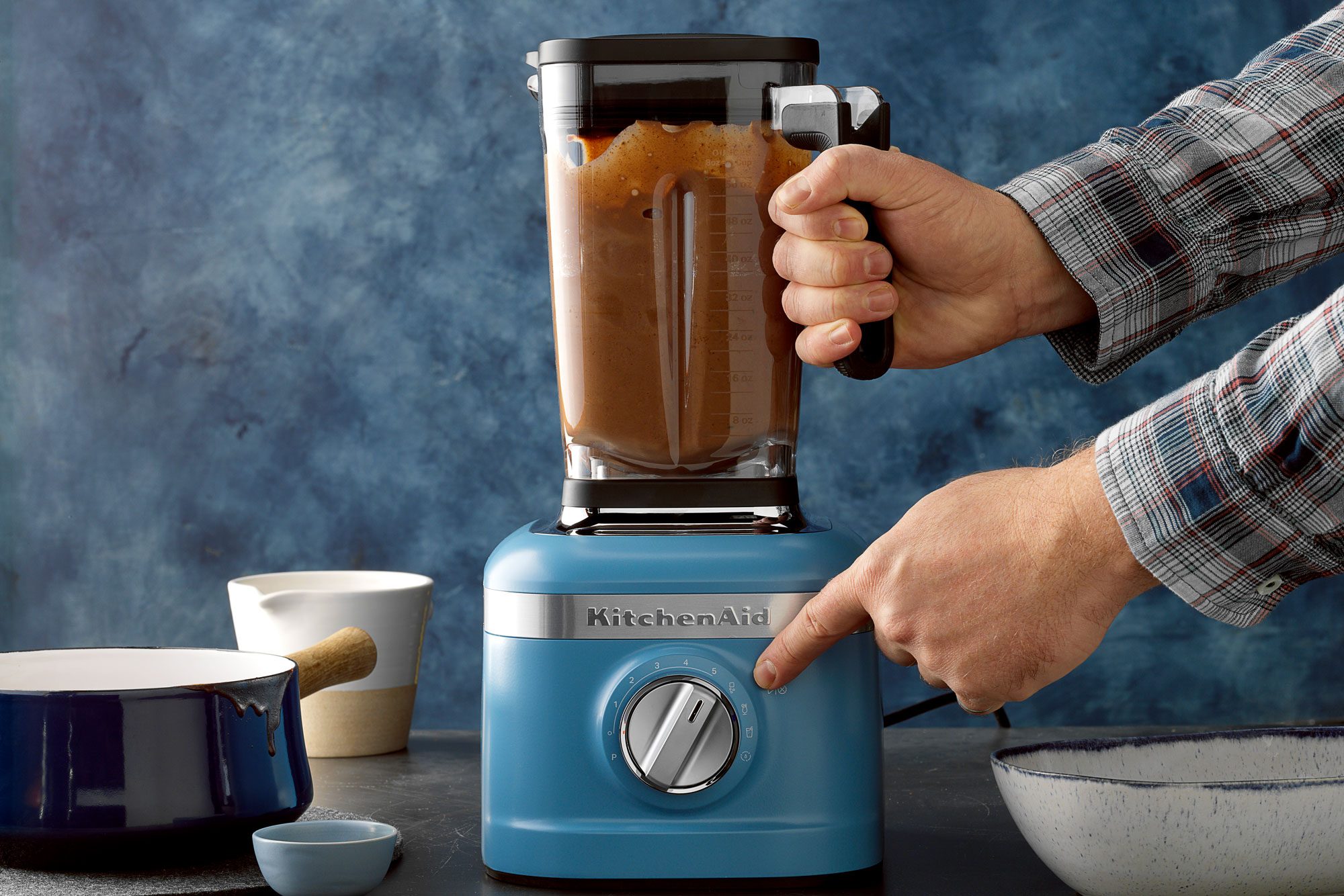A person is using a blue KitchenAid blender, filled with a chocolate mixture