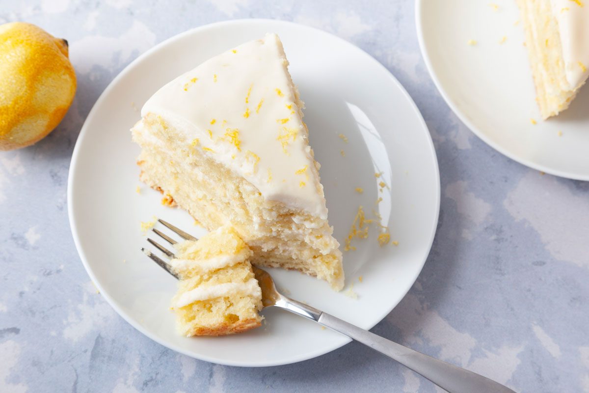 Single servings of Taste of Home Italian Lemon Cream Cake on white pates, garnished with lemon zest, on a light blue surface, zested lemon peaking in