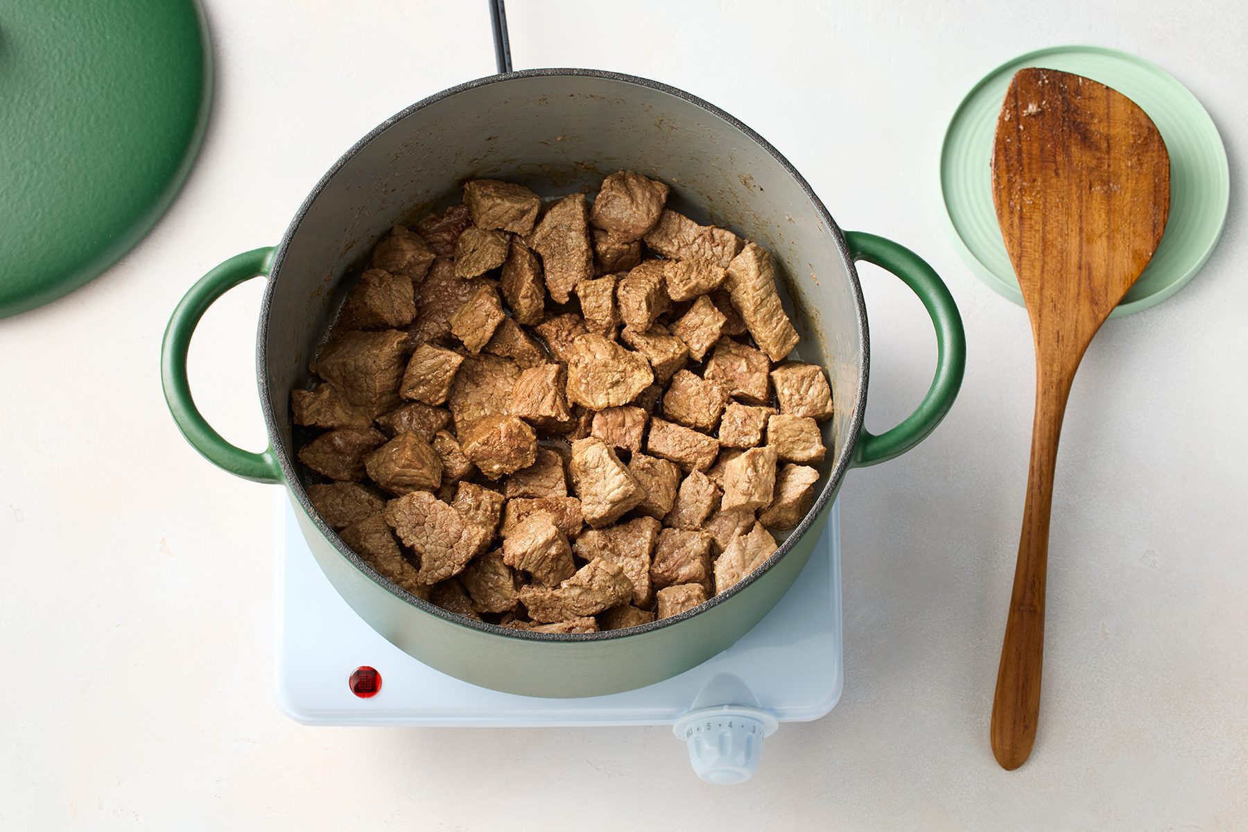 A green pot filled with browned chunks of meat is placed on a white electric stovetop. 