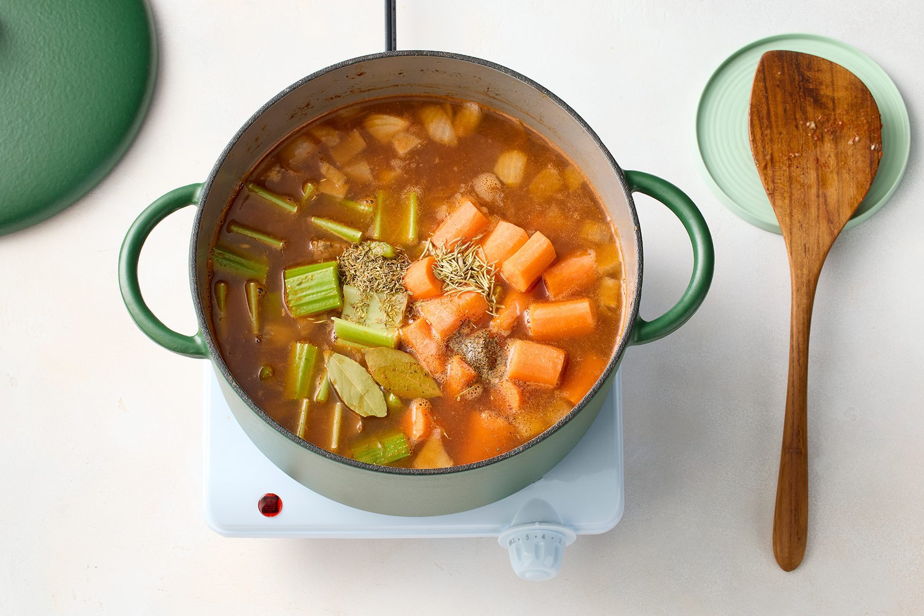 A green pot containing a vegetable stew with chunks of carrots, celery, onions, and spices is simmering on a hot plate. 