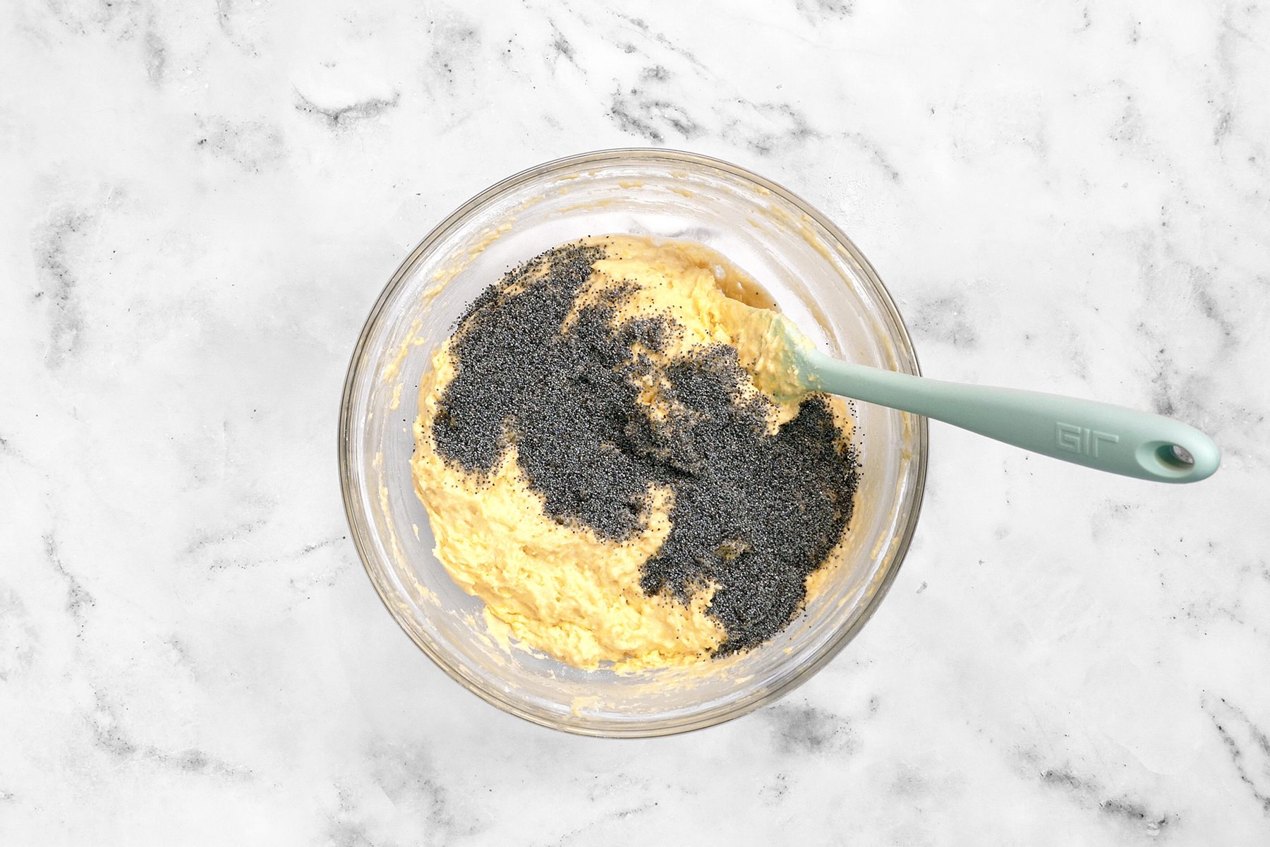 A clear glass mixing bowl filled with a yellow batter and topped with black poppy seeds. 