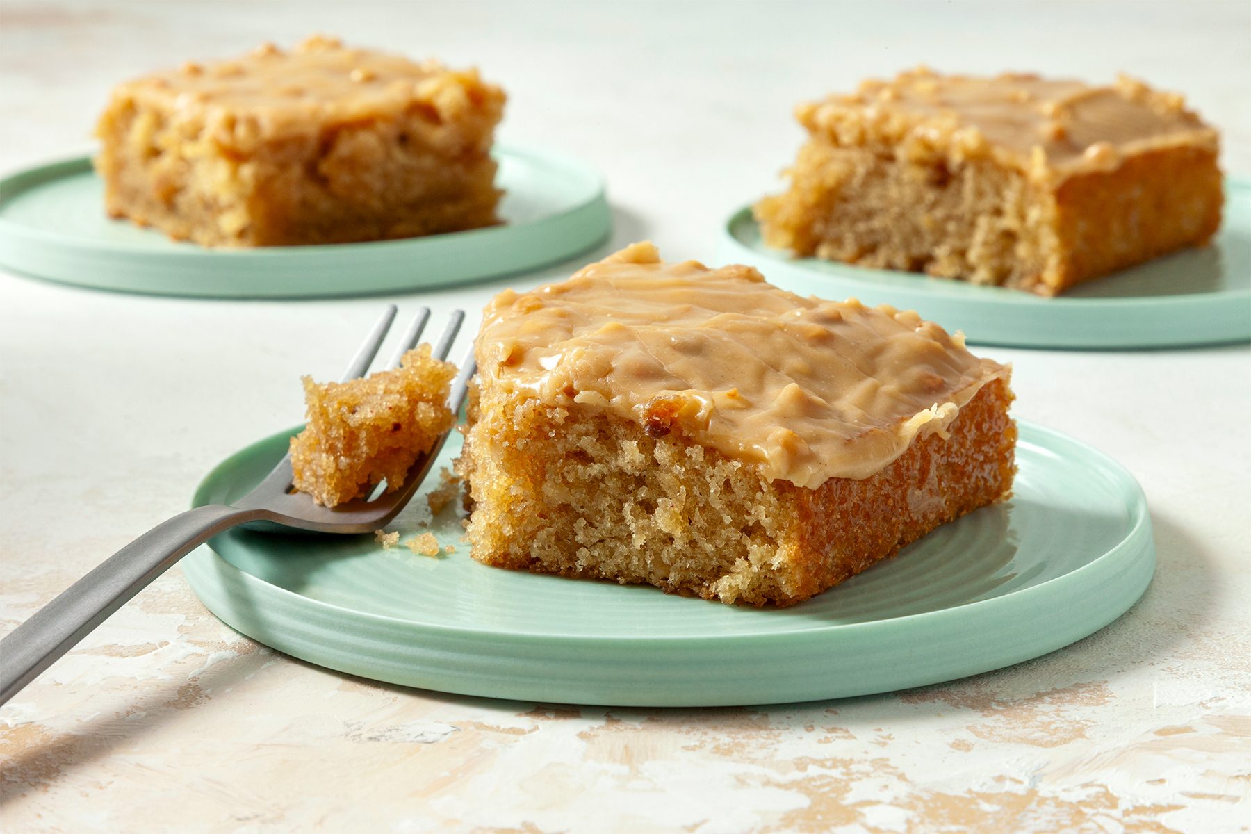 close shot; white textured background; Peanut Butter Sheet Cake cut into square pieces served on three small plates with silver fork;