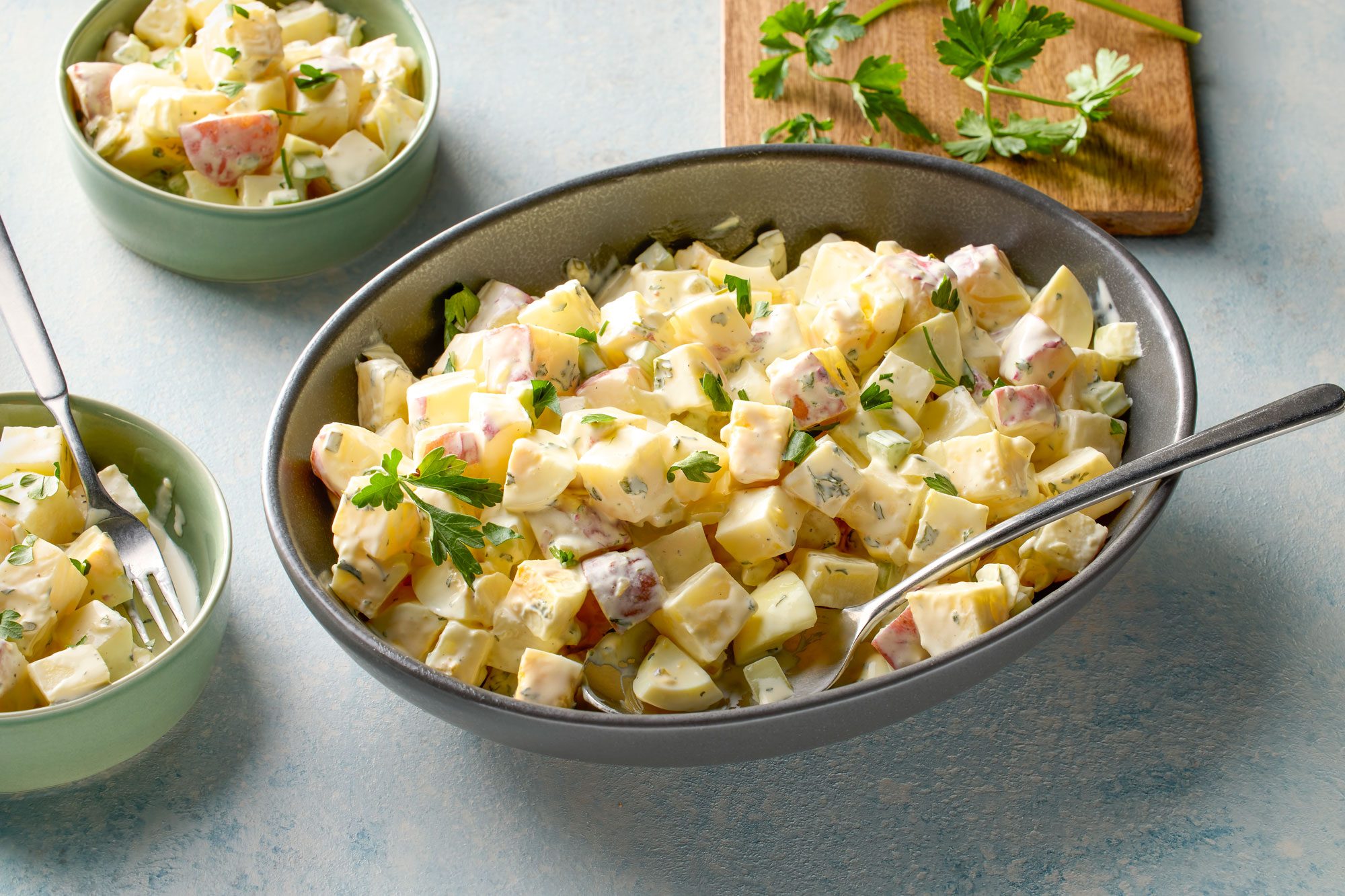 Top view shot of Red Potato and Egg Salad; in a large bowl; with serving spoon; served in two bowls with fork; wooden chopping board; celery; blue and white texture background;