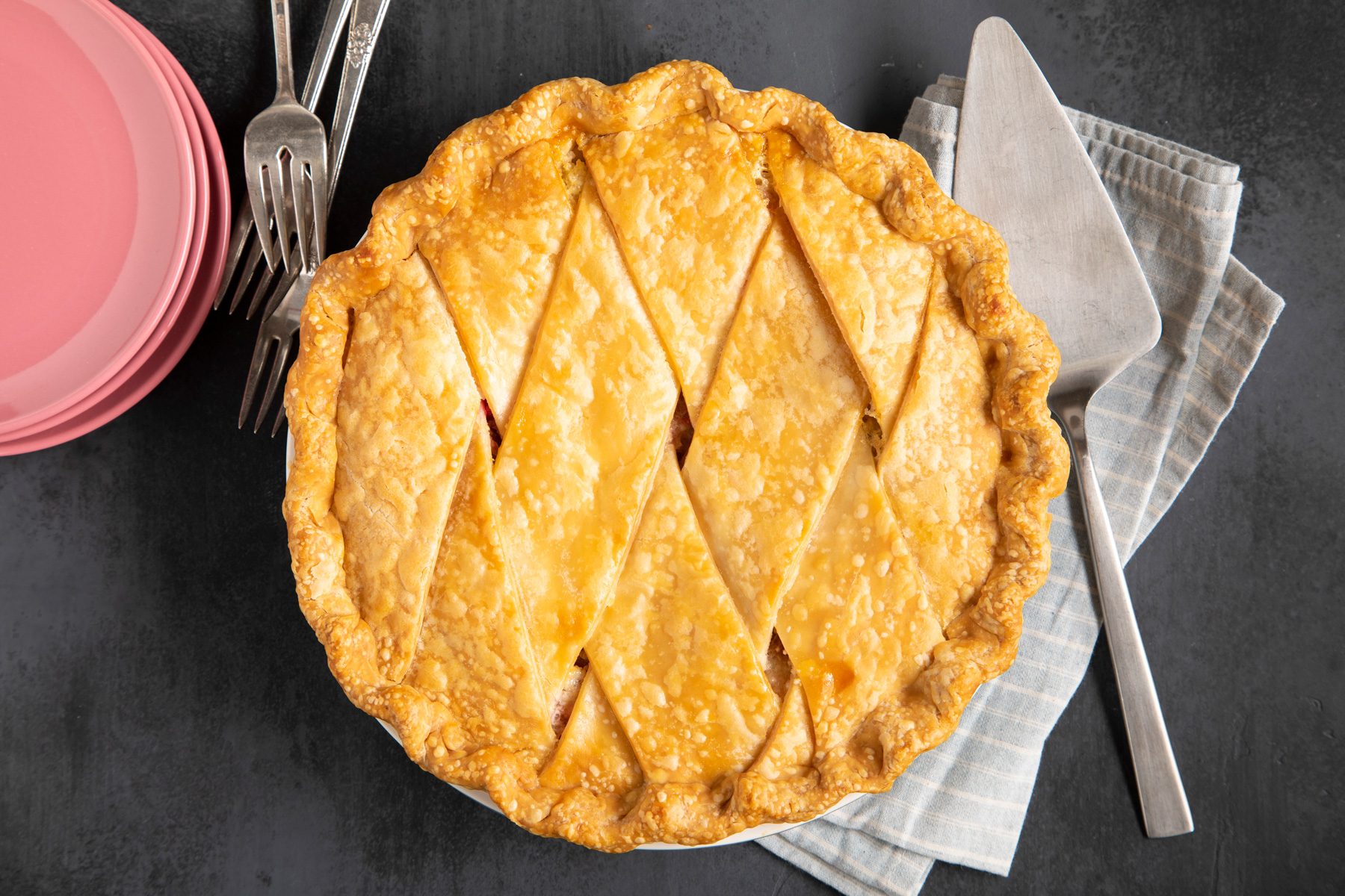 An overhead view of a freshly baked Rhubarb Custard Pie with plates, forks, and a pie server.