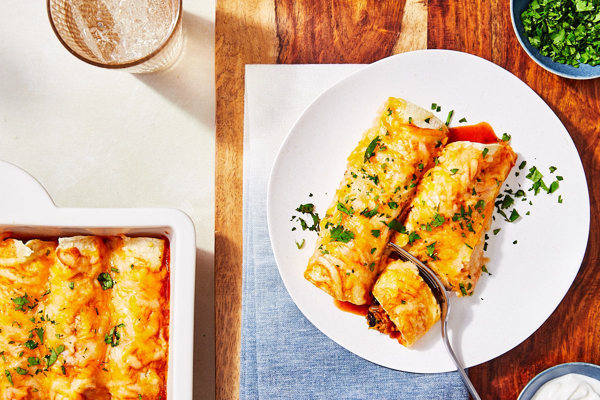 Smoky Sweet Potato And Black Bean Enchiladas Hero- 2 enchiladas plated and topped with cilantro, fork cutting into one enchilada
