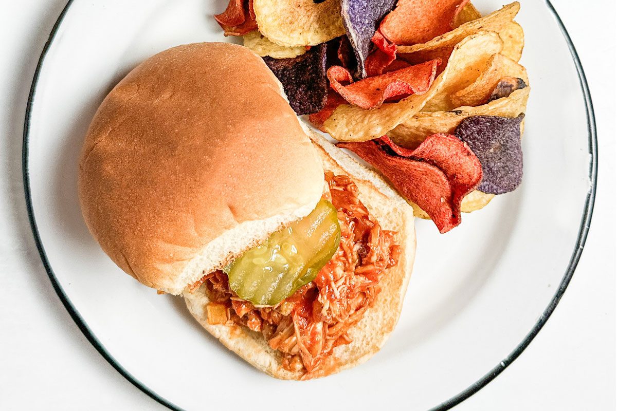 Taste of Home Barbecue Chicken Sandwich with chips on enamel plates next to bowls of chips and pickles