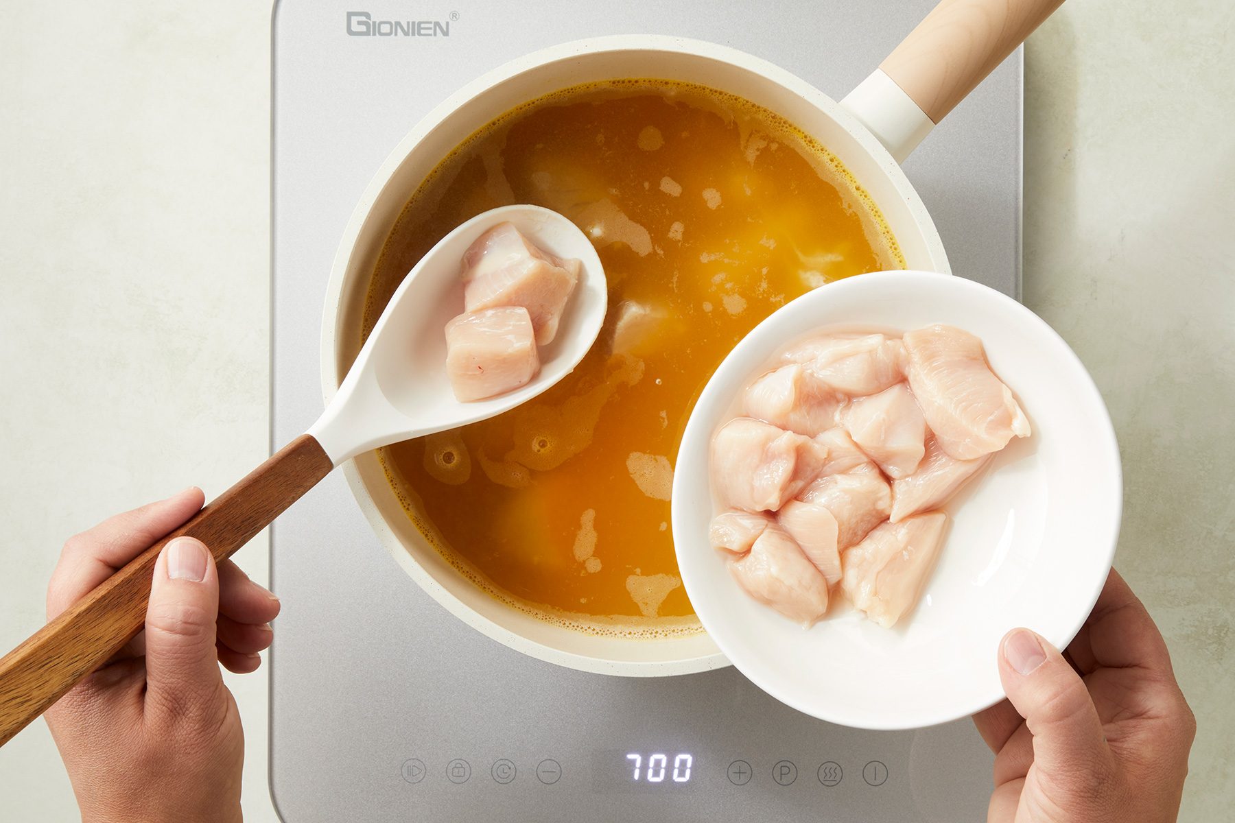 A person is adding cubed pieces of raw chicken from a white bowl into a pot of soup on a stove. The soup appears to be a yellowish broth. The spoon used to transfer the chicken is white with a wooden handle.