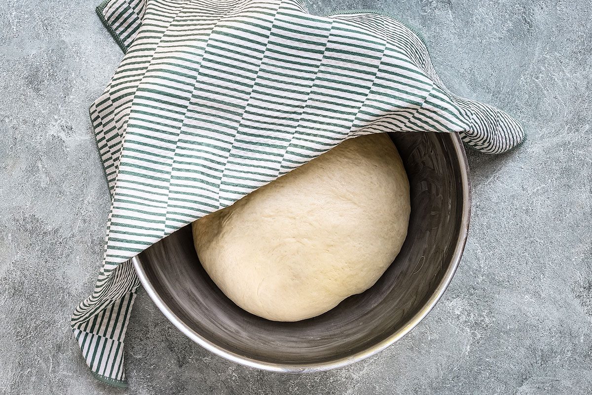 Dough for Taste of Home cinnamon roll focaccia ready to rise in a bowl with cloth covering.