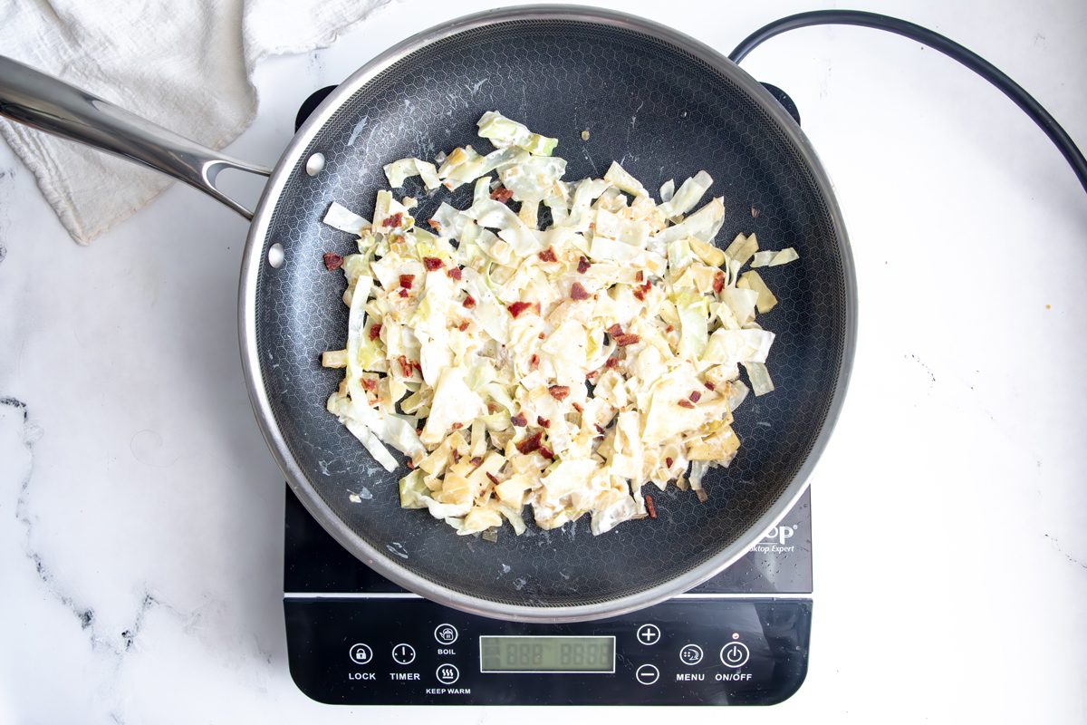 Cooked cabbage and apple topped with bacon in a Hexclad pan with bacon grease for Taste of home creamed cabbage on an induction cooktop on a marble surface.