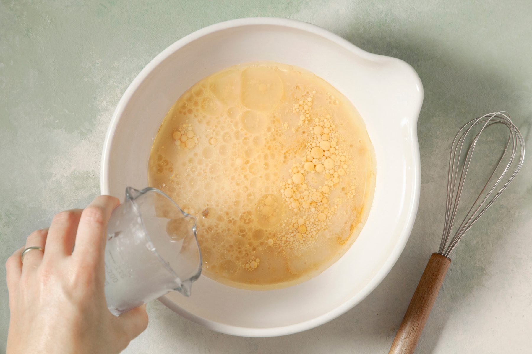 overhead shot; light green textured background; In another bowl, pouring water from a measuring cup into the bowl, combined with almond milk, oil and vanilla until well blended;