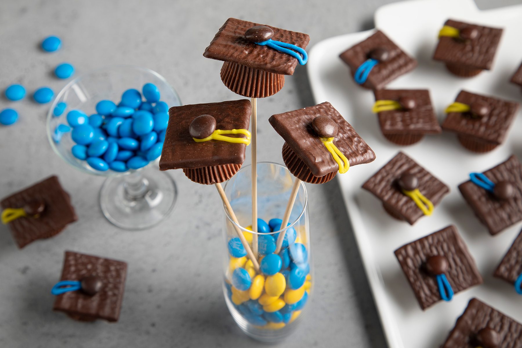 Chocolate graduation cap treats on sticks, with mini peanut butter cups as bases, chocolate squares as caps, and colored candy tassels, displayed in a glass filled with blue and yellow candy-coated chocolates. A dish of similar candies is in the background.