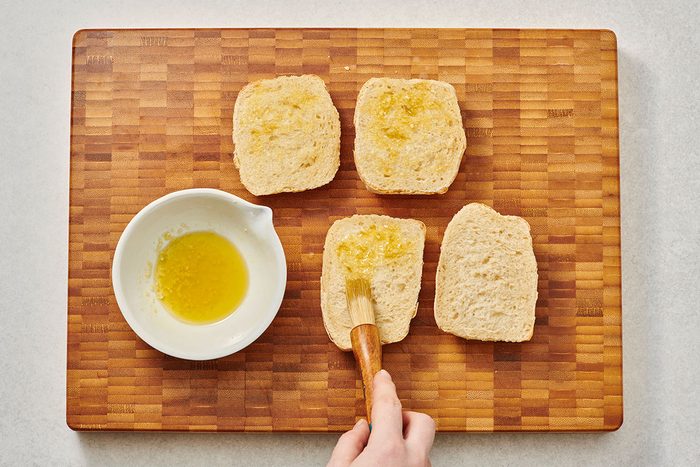 Brushing ciabatta roll slices with olive oil and garlic paste on a cutting board
