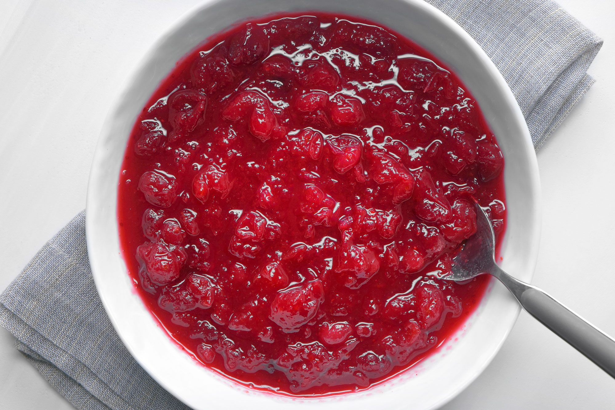 overhead shot; A white bowl is filled with a vibrant red cranberry sauce, placed over kitchen towel