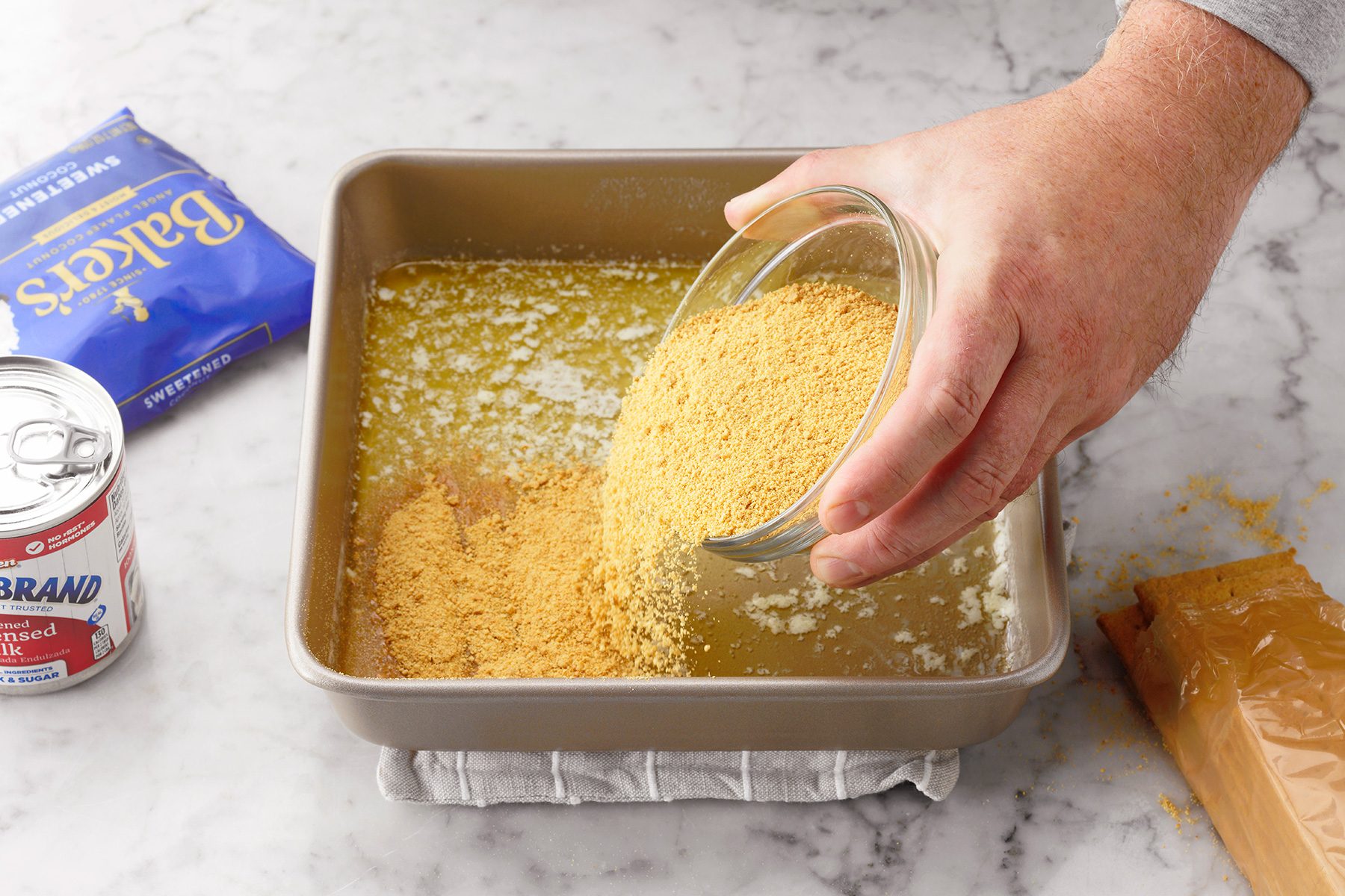A close-up view of a person pouring crushed graham crackers from a glass bowl into a square baking pan with melted butter. A can of sweetened condensed milk and a package of Baker's chocolate are positioned on the marble countertop nearby.