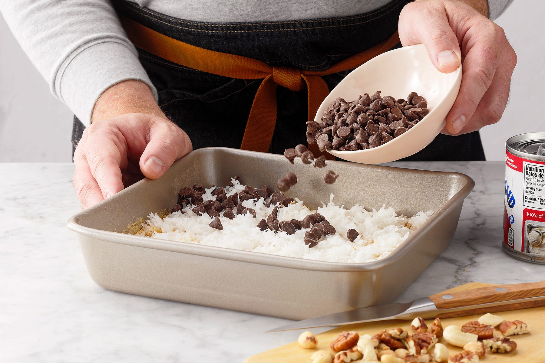 A person wearing a gray shirt and black apron is sprinkling chocolate chips from a small bowl onto a baking pan filled with a white ingredient, possibly shredded coconut. The countertop also holds chopped nuts and a can of condensed milk.
