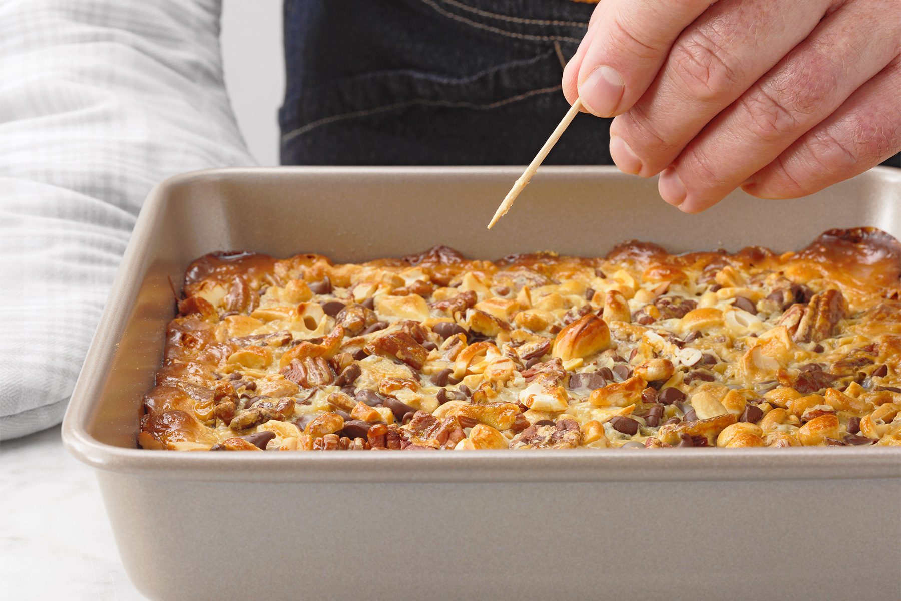 A hand holding a toothpick tests the doneness of a dessert that appears to be a baked bar or cake with a golden-brown top and visible nuts, in a rectangular baking dish.