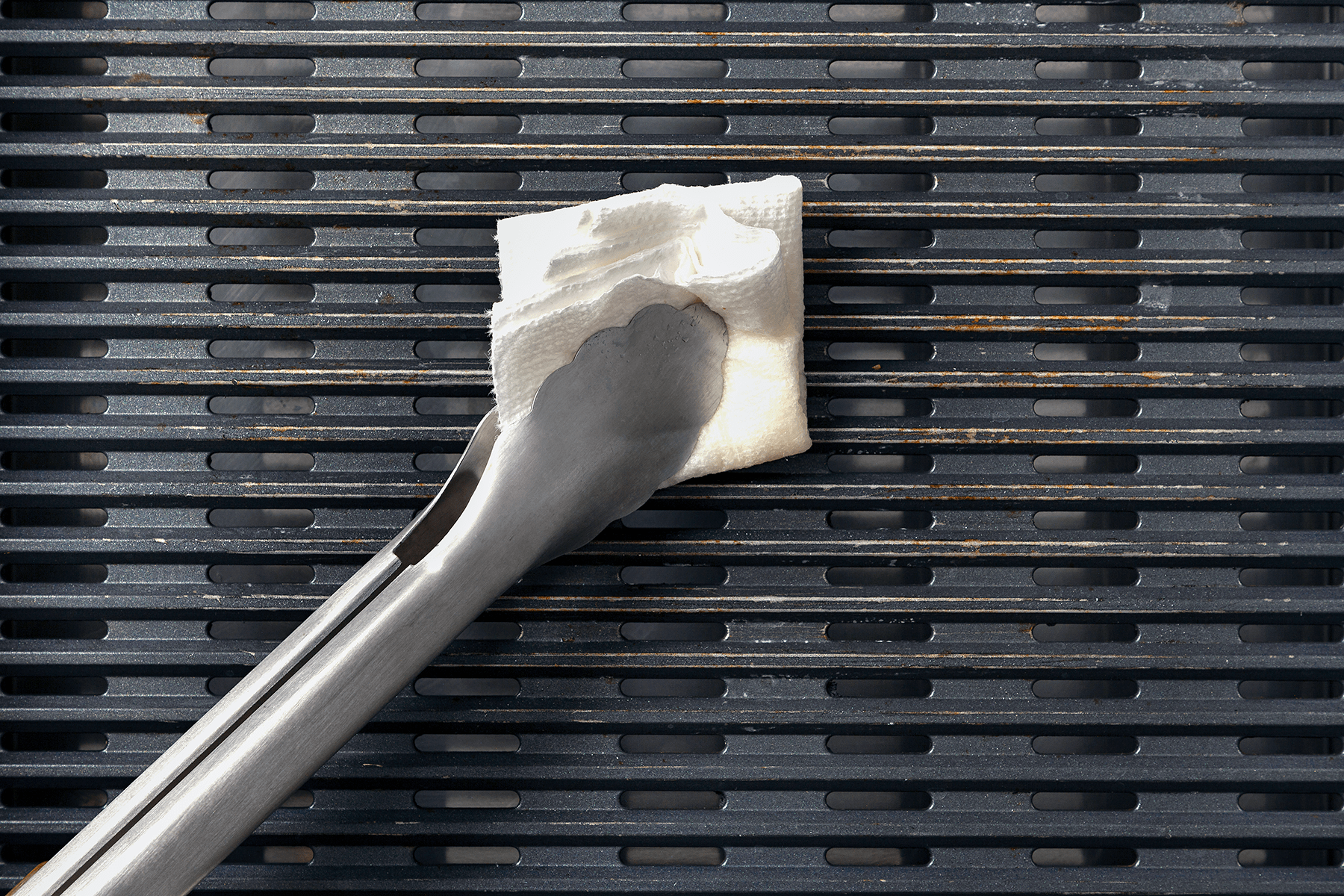 Metal tongs holding a piece of folded white paper towel are being used to clean the grates of a barbecue grill. The grill grates are black with a slightly worn appearance.