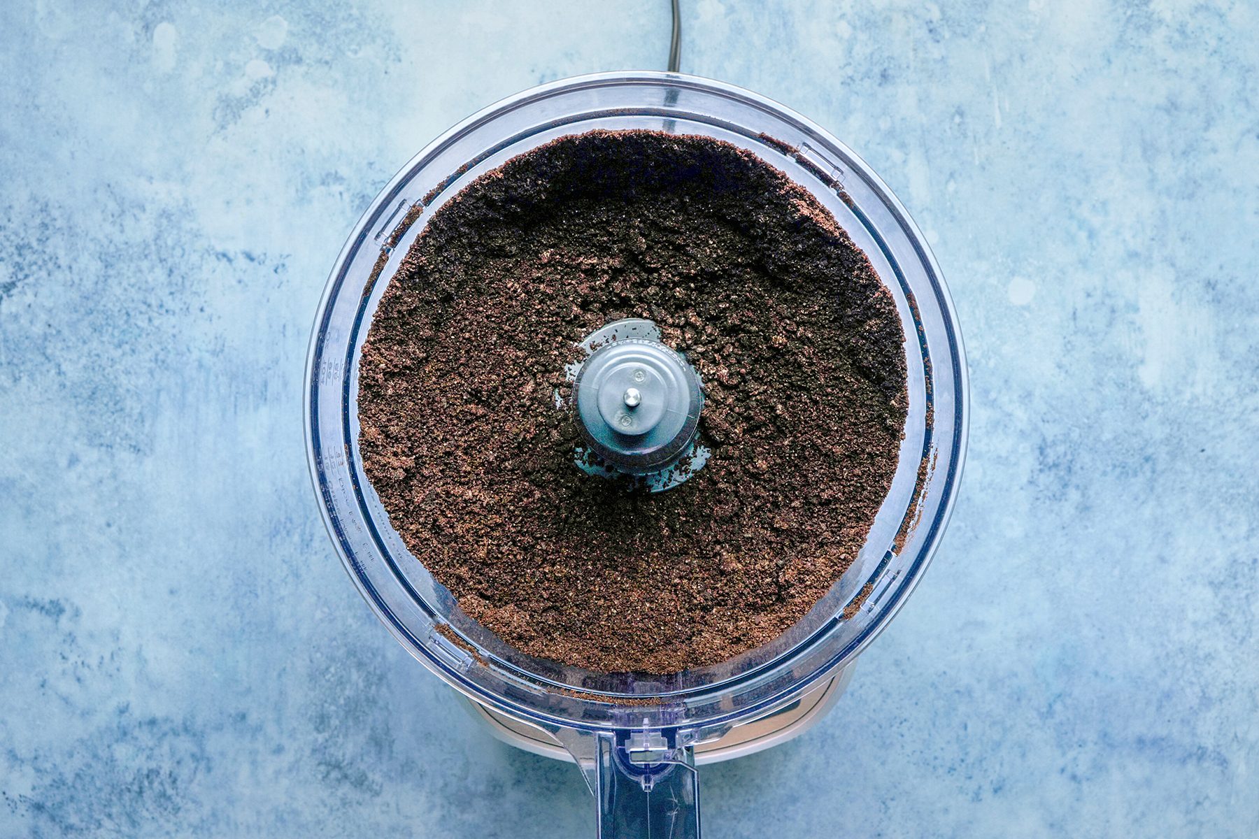 Top view of a food processor containing finely ground chocolate cookie crumbs. The background is a light blue, textured surface.