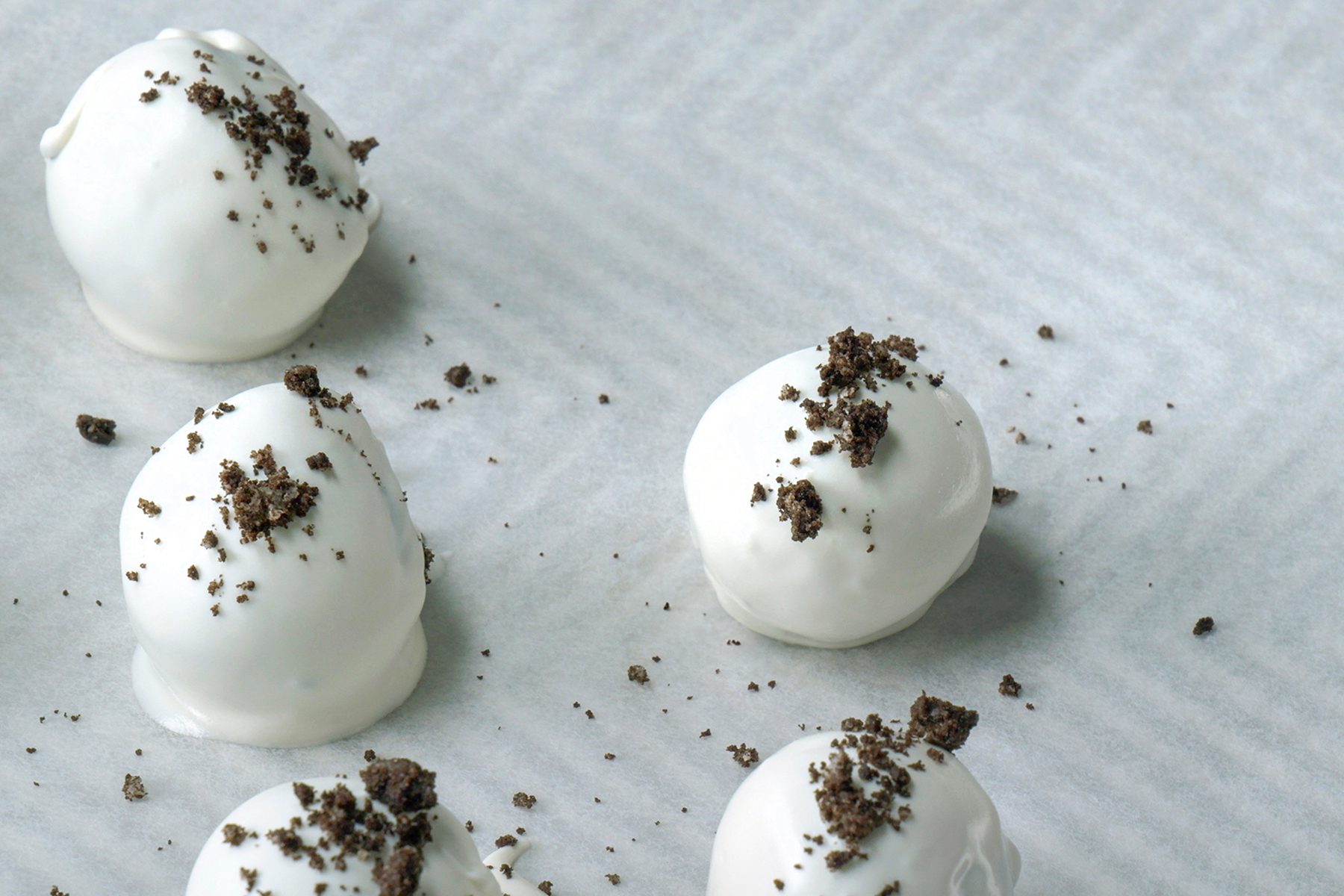 Six white chocolate-covered truffles are placed on a sheet of parchment paper. Each truffle is sprinkled with small, dark chocolate crumbs on top.