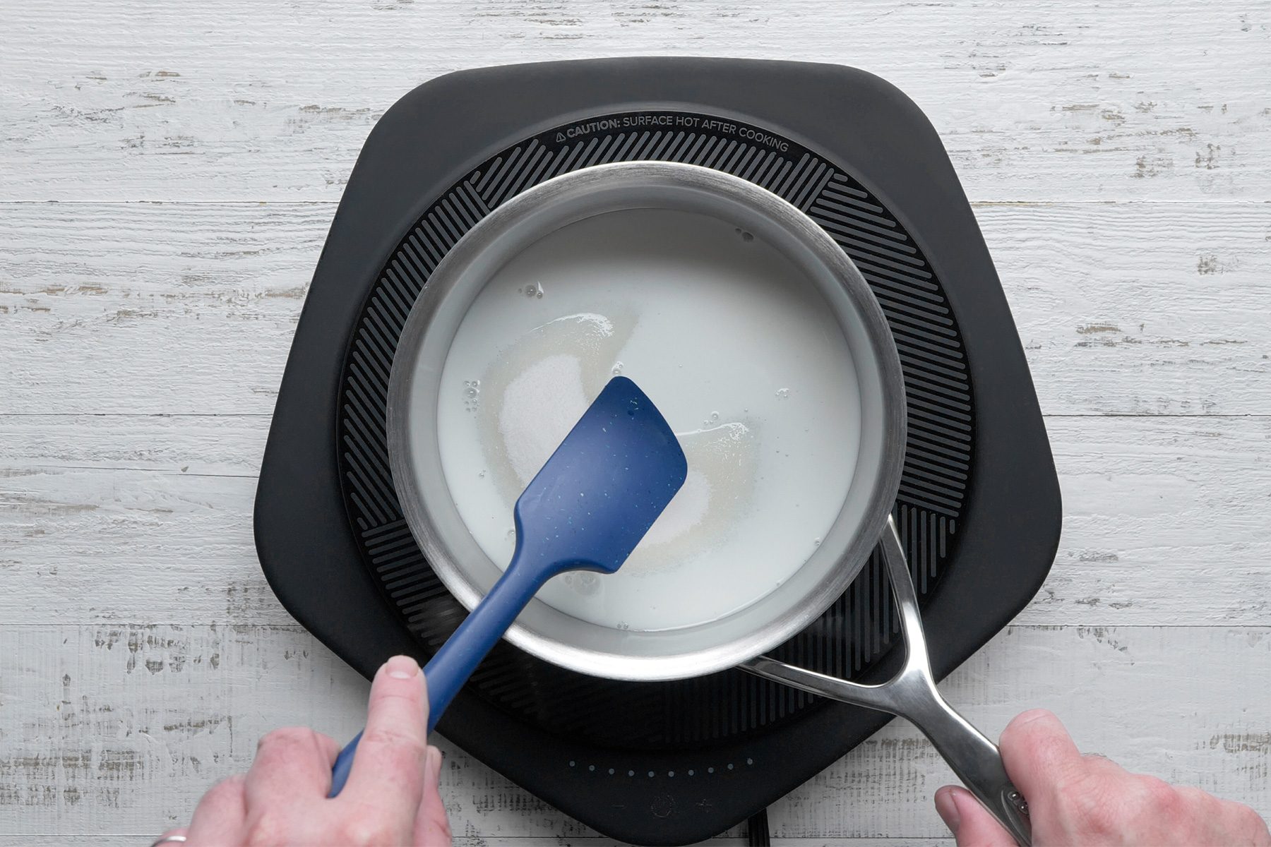 Overhead shot of combined sugar and milk in aheavy saucepan; bring to boil; spatula; induction; white wooden background;