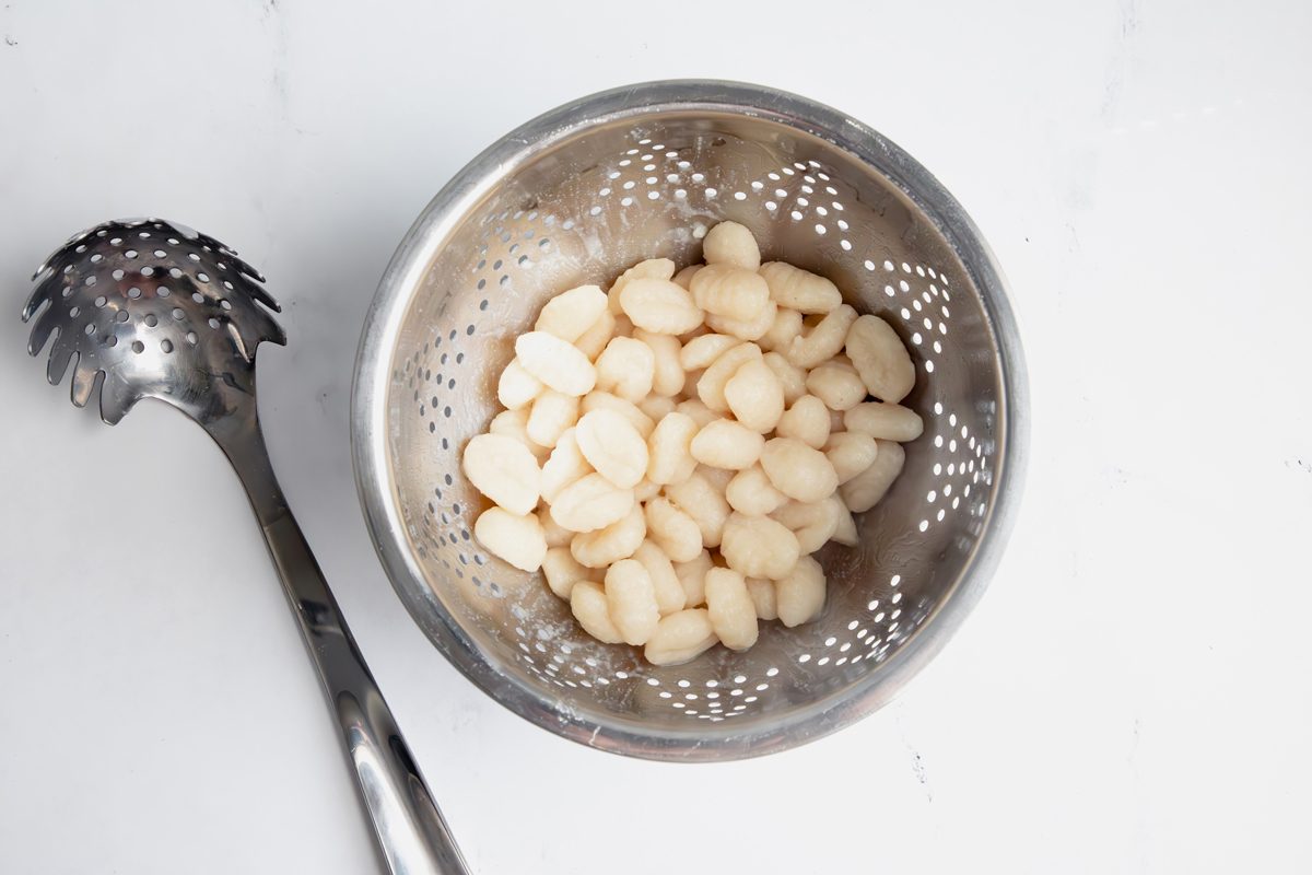 Cooked gnocchi in a metal colander for Taste of Home Pesto Gnocchi with a pasta fork on a marble surface.