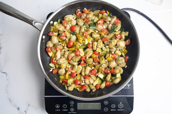 Peppers, zucchini, gnocchi, and tomatoes in a skillet on an induction cooktop for Taste of Home Pesto Gnocchi on a marble surface.