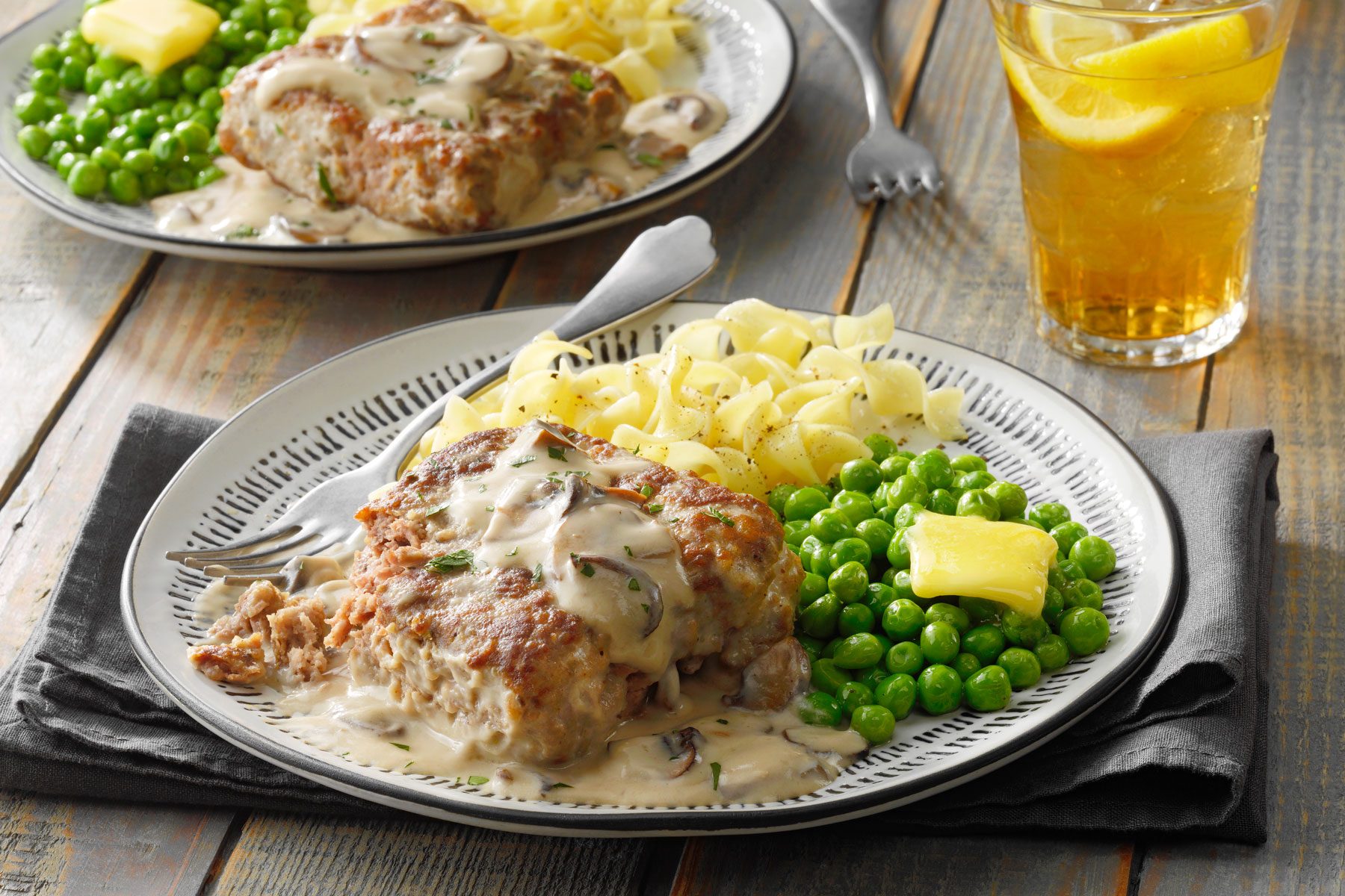 Two plates of hearty meals with sliced pork chops covered in creamy mushroom sauce, sides of green peas, and buttered egg noodles. Each plate is garnished neatly. In the background is an ice-cold drink with lemon slices. The meal is set on a wooden table.