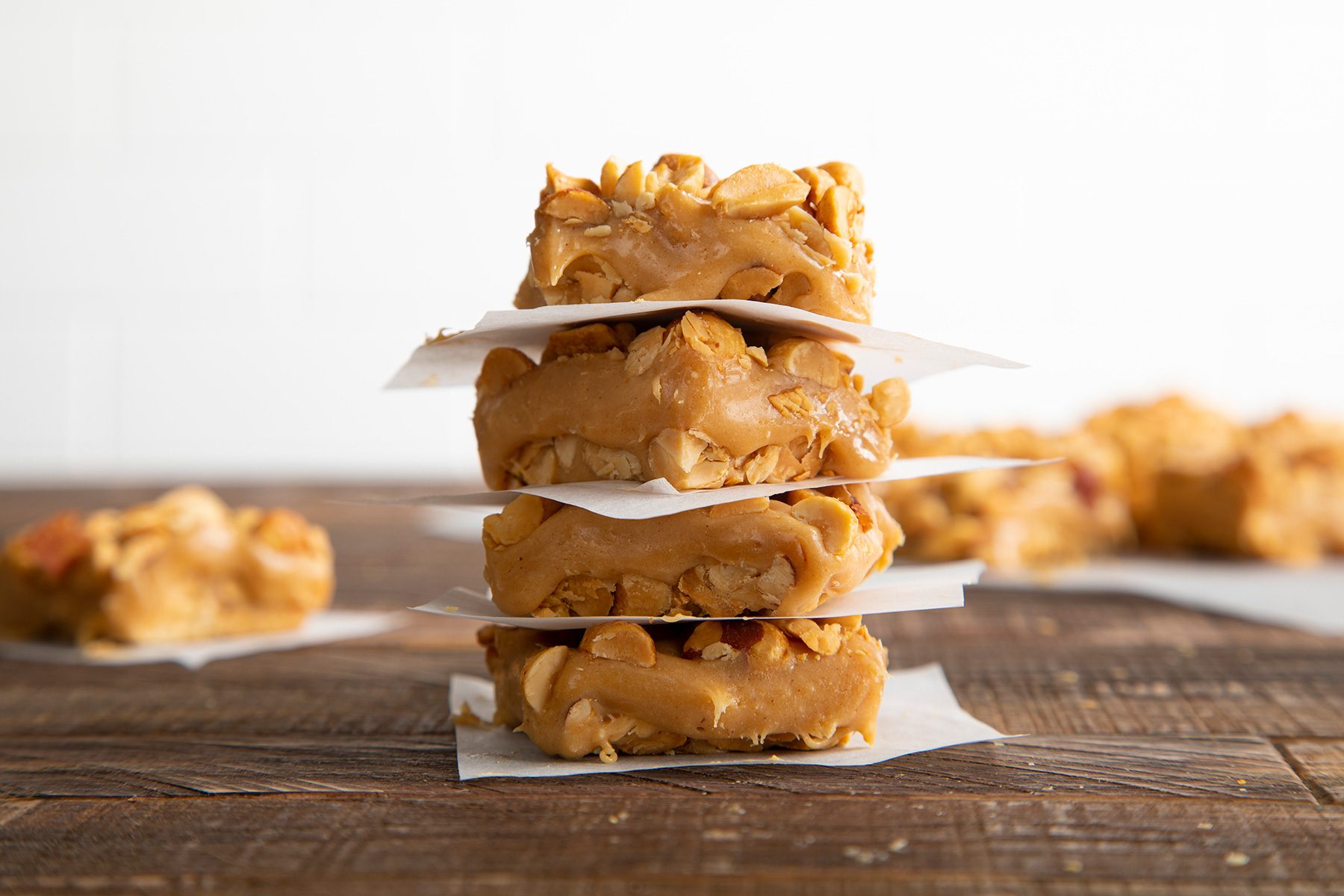 A close-up of four peanut butter bars stacked on top of each other, separated by pieces of parchment paper. The bars contain visible peanuts and have a rich, creamy texture. They are placed on a rustic wooden surface with a blurred background.